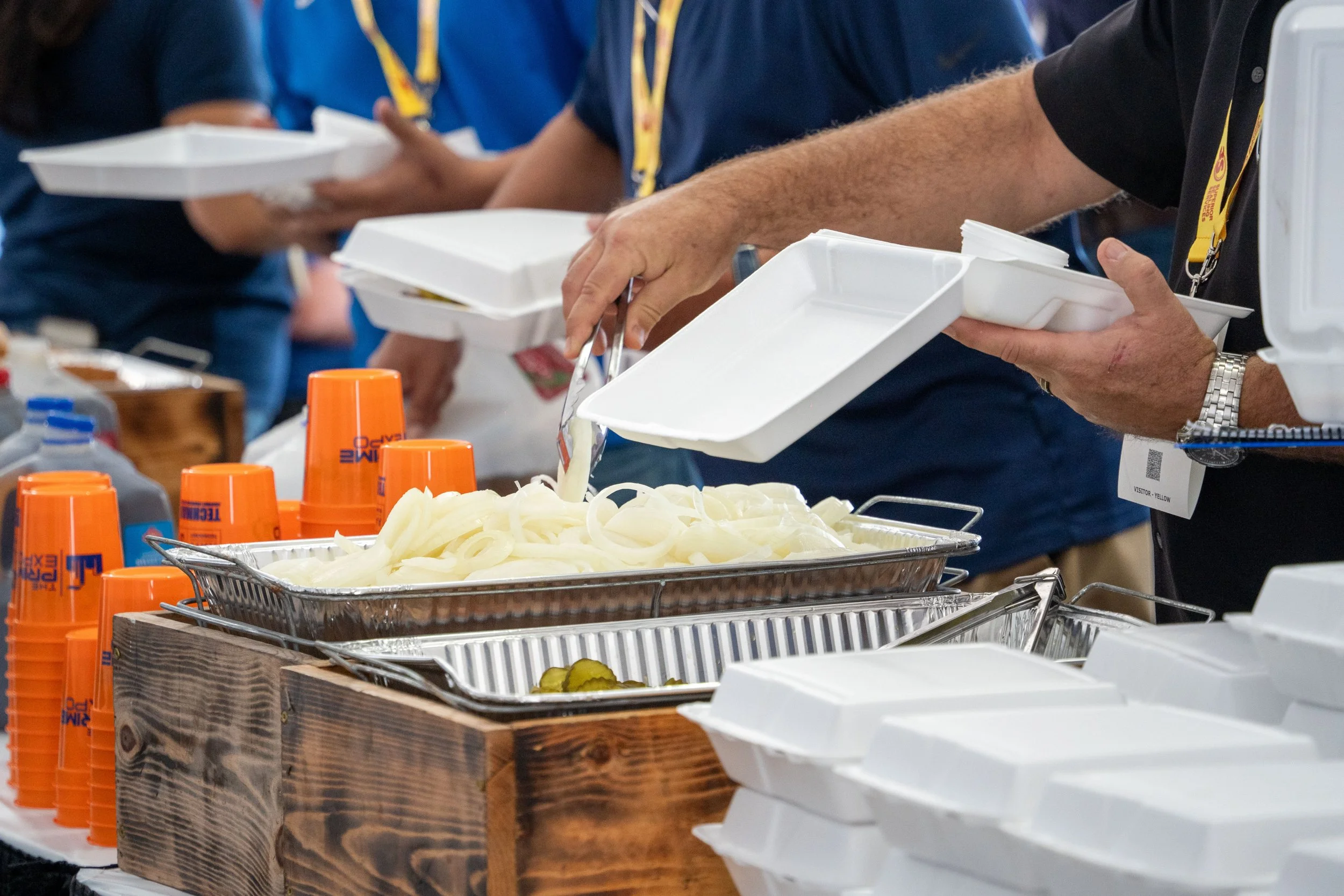 People serving themselves food at a buffet with trays of onions and pickles, disposable cups, and styrofoam takeout containers.