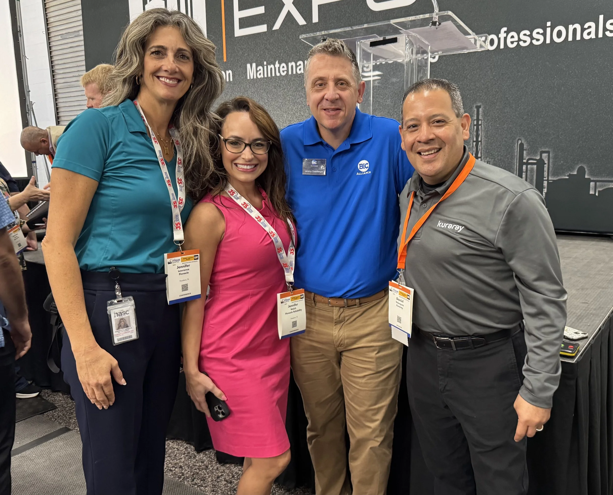 Four professionals, two women and two men, standing together at a conference, smiling at the camera. They are wearing conference badges and lanyards, with a technology or industrial background behind them.