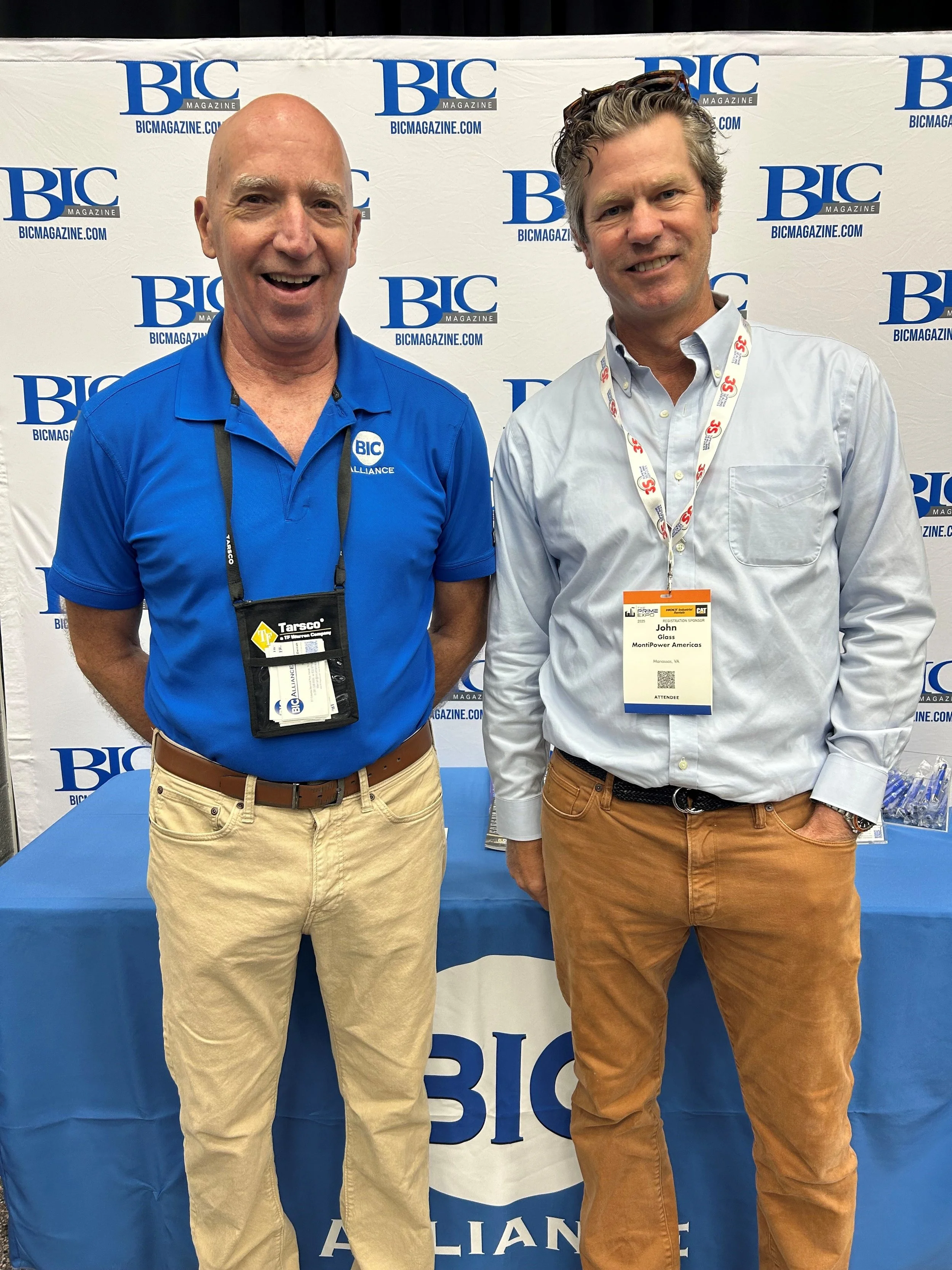 Two men standing in front of a BIC magazine backdrop at a conference or event. The man on the left is wearing a blue polo shirt with the BIC logo, beige pants, and has a lanyard with a badge. The man on the right is wearing a light blue button-up shi