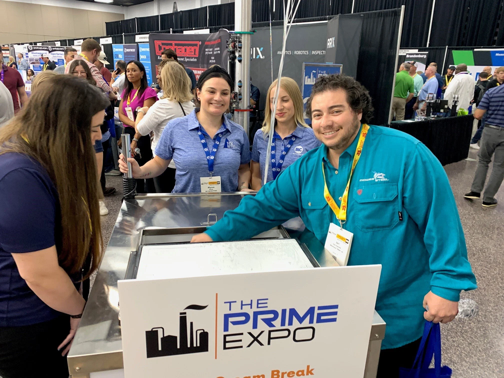 Man smiling at a table at The Prime Expo with three women behind him, all wearing blue shirts with logos, at a busy exhibition hall.