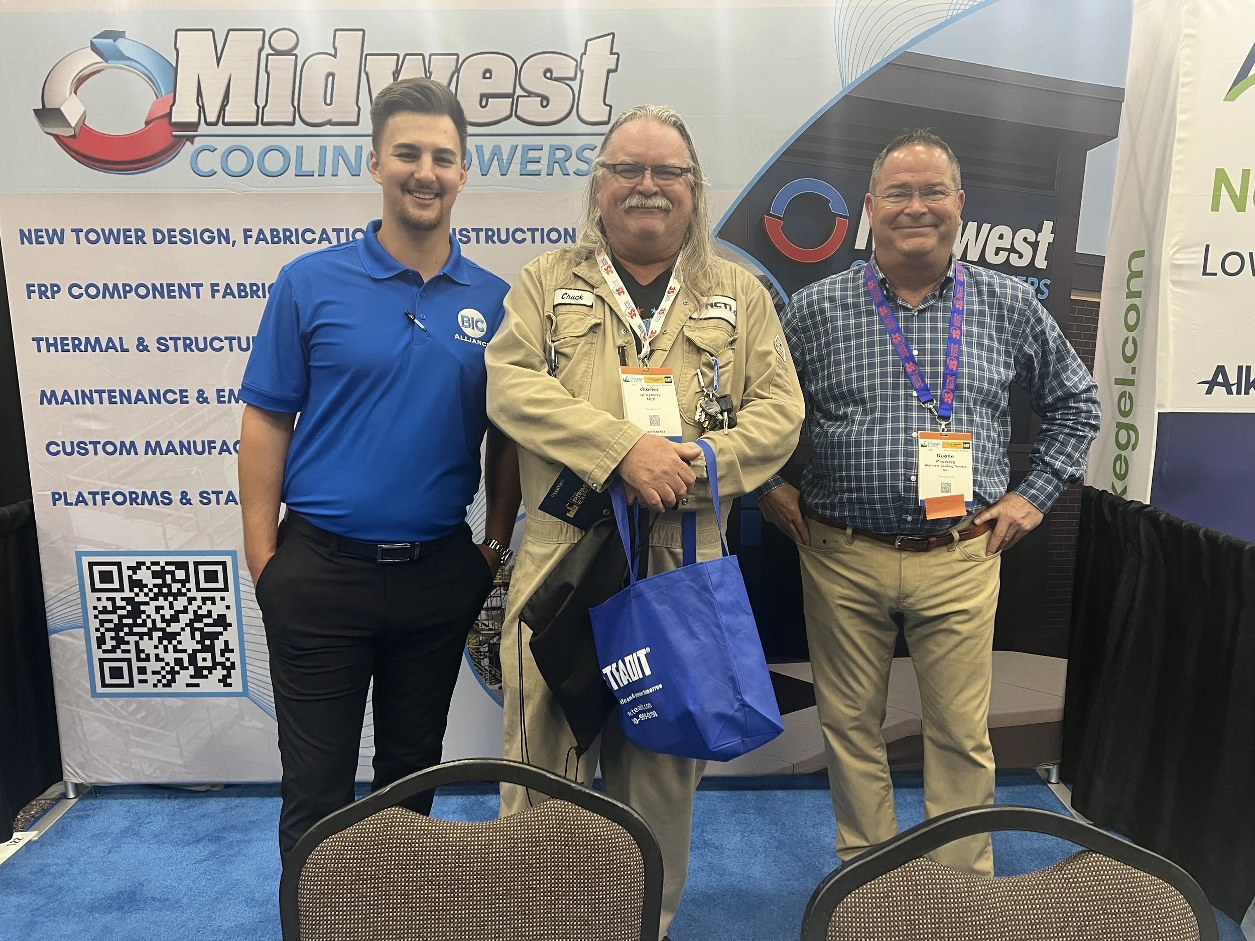 Three men standing in front of a trade show booth with the MidWest Cooling Towers logo, smiling for the camera. The booth has informational text about tower design, fabrication, and construction, with a QR code at the bottom. The man in the center we