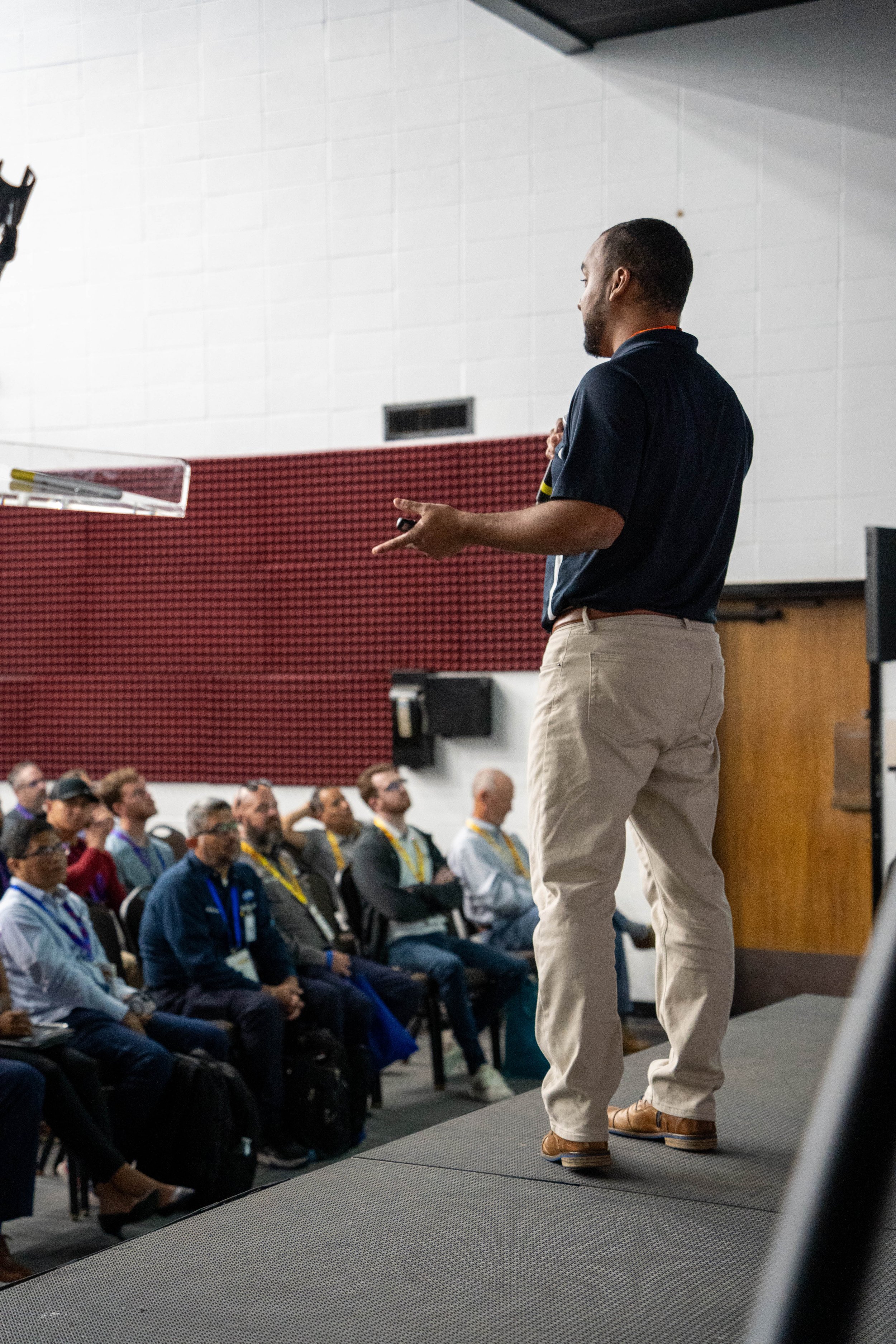 A man stands on a stage gesturing to an audience in a conference room.