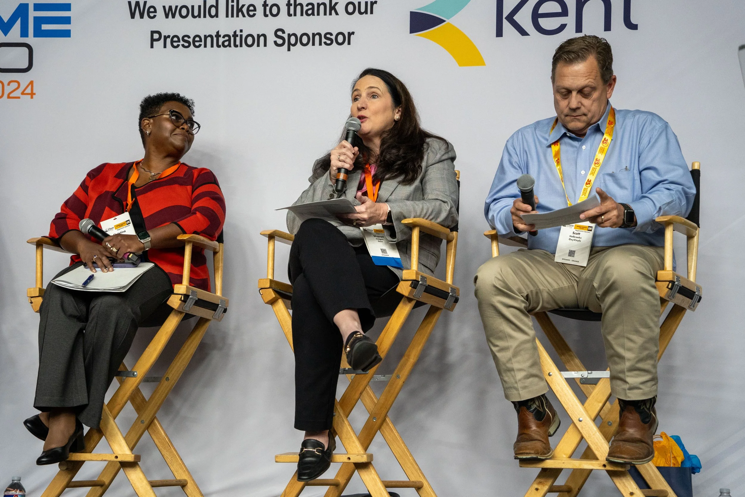 Three people are seated on a panel at a conference, each holding a microphone. They are sitting on wooden director's chairs against a backdrop with text and logos. The woman on the left is wearing glasses and a red and black striped top, the woman in