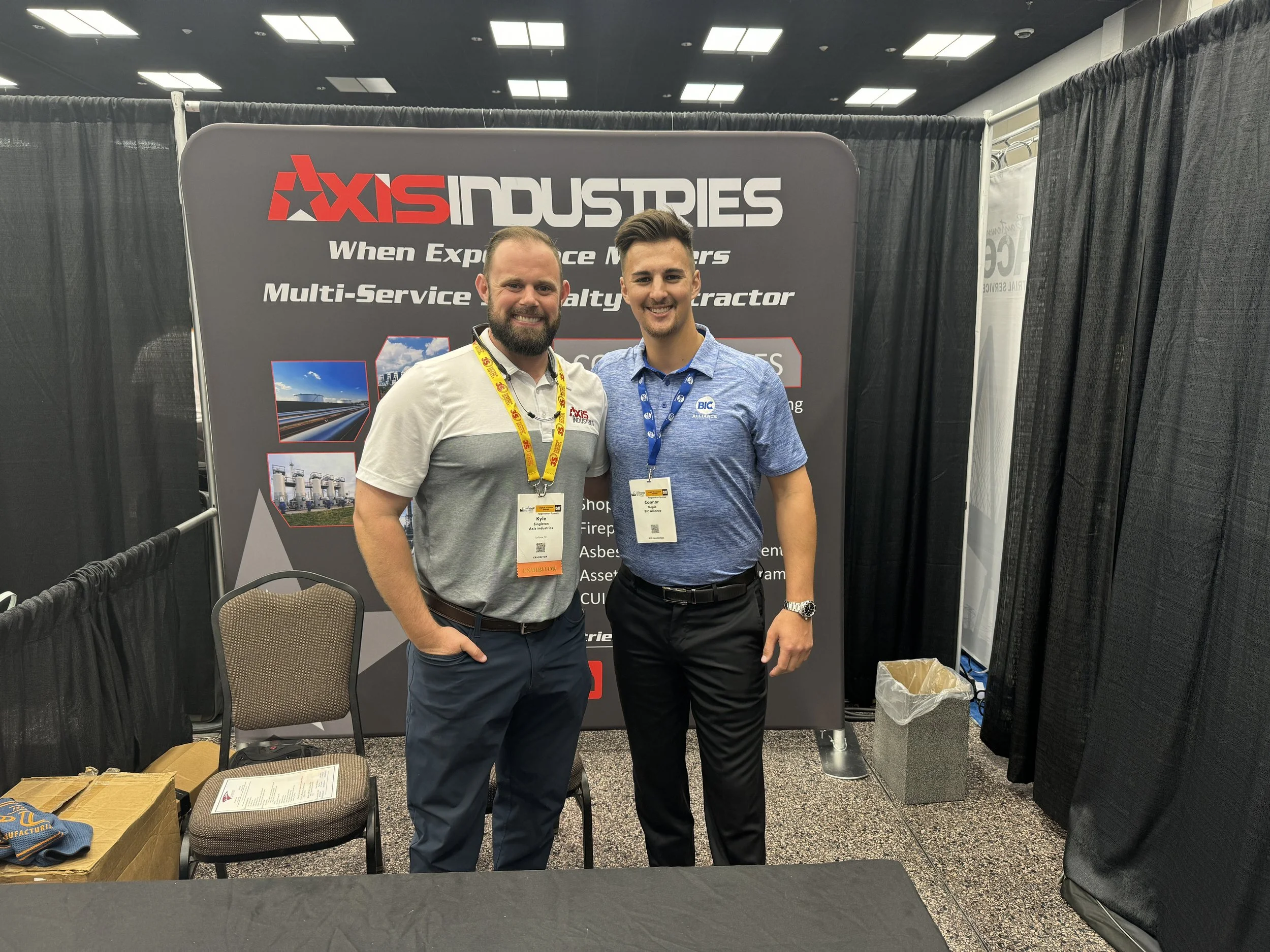 Two men standing in front of an Axis Industries booth at a trade show, smiling and posing for the camera. One man is wearing a white and gray polo shirt with a yellow lanyard, and the other is wearing a blue polo shirt with a blue lanyard. Behind the