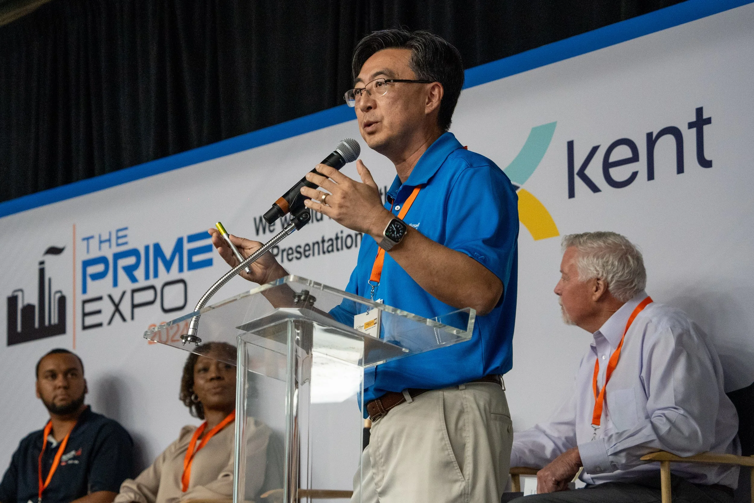 A man speaking at a podium during a conference with a banner that reads 'The Prime Expo' in the background, wearing glasses, a blue shirt, and an orange lanyard. Two other people are seated behind him, one in a white shirt and the other in a beige ja