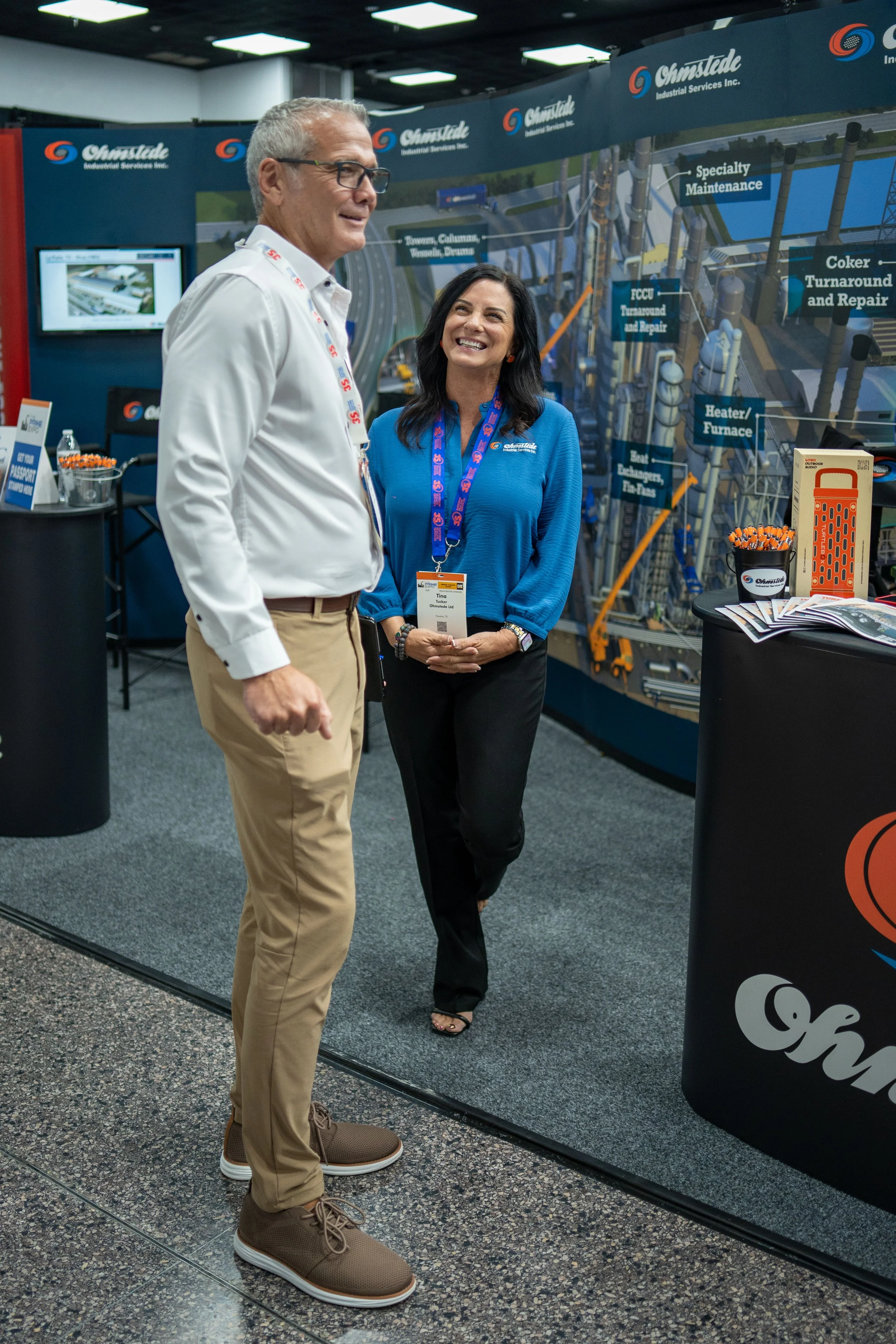Two people, a man and a woman, are standing and talking at an industrial services booth at a trade show. The man is wearing glasses, a white shirt, khaki pants, and brown shoes. The woman is dressed in a blue blouse and black pants. Behind them is a 