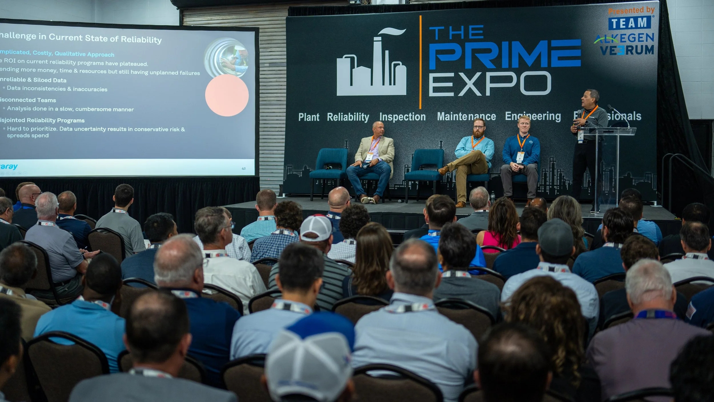 A conference panel titled 'The Prime Expo' with four male speakers on stage, one standing at a clear podium speaking to an audience, three seated on stage with blue chairs. Behind them is a large screen displaying a presentation slide with text about