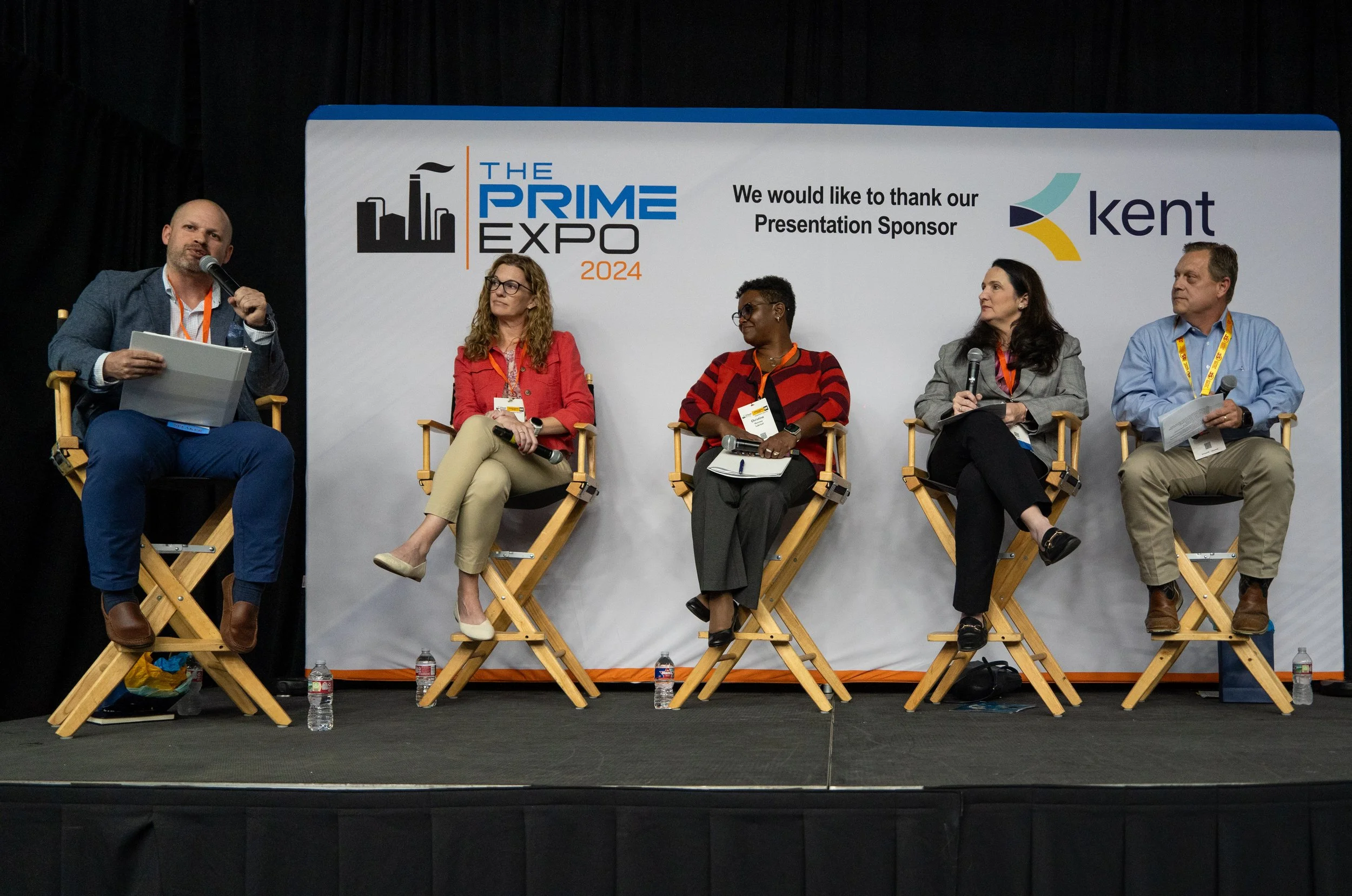 Panel discussion at The Prime Expo 2024 with five speakers seated on stage in director chairs, microphones in hand, and a large backdrop displaying event branding and sponsor information.