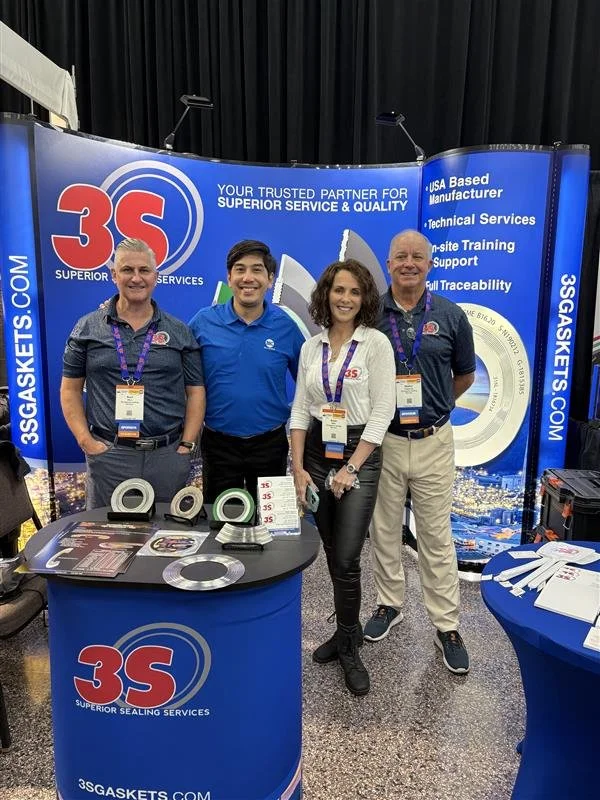 Four individuals standing behind a booth at a trade show for 3S Gaskets, a company specializing in sealing services. The booth has banners with the company logo and information about their services, including being a USA-based manufacturer with techn