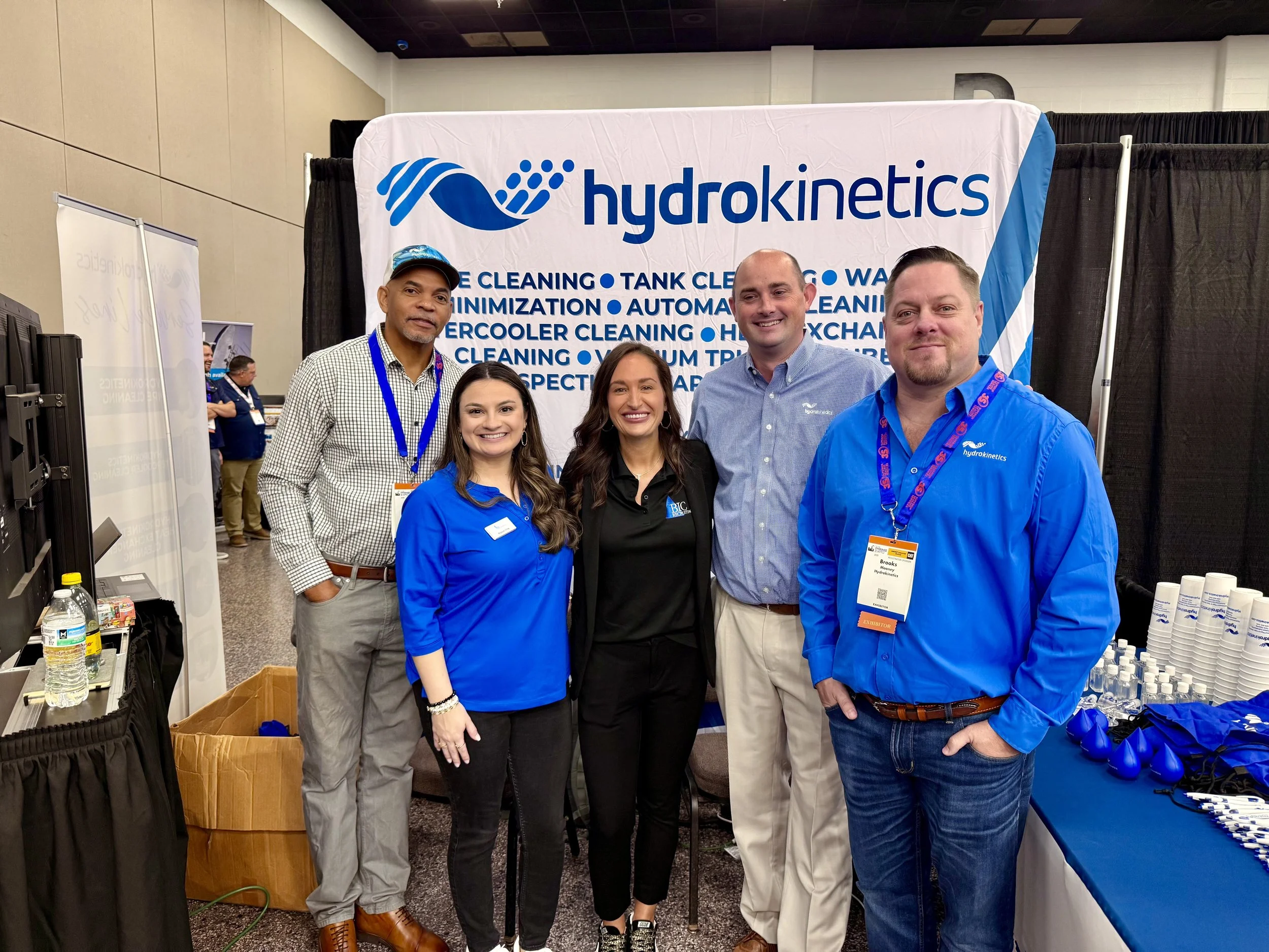 Group of five people standing in front of a Hydrokinetics booth at a trade show, smiling, with promotional materials on a table to the right.