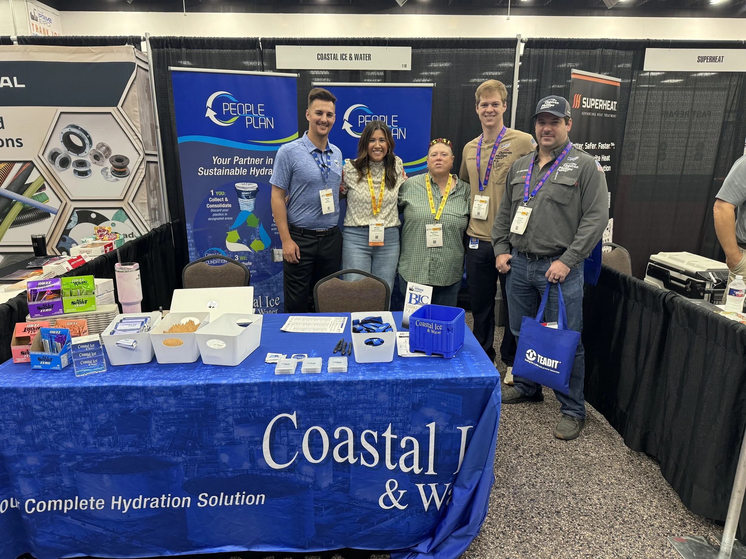 Group of five people standing behind a booth at a trade show, with two blue banners for People Plan and Coastal Ice & Water, and a table covered with a blue tablecloth displaying promotional materials.