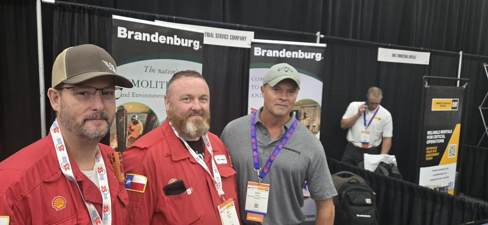 Three men posing at a trade show booth with banners for Brandenburg, a trial service company, in the background.