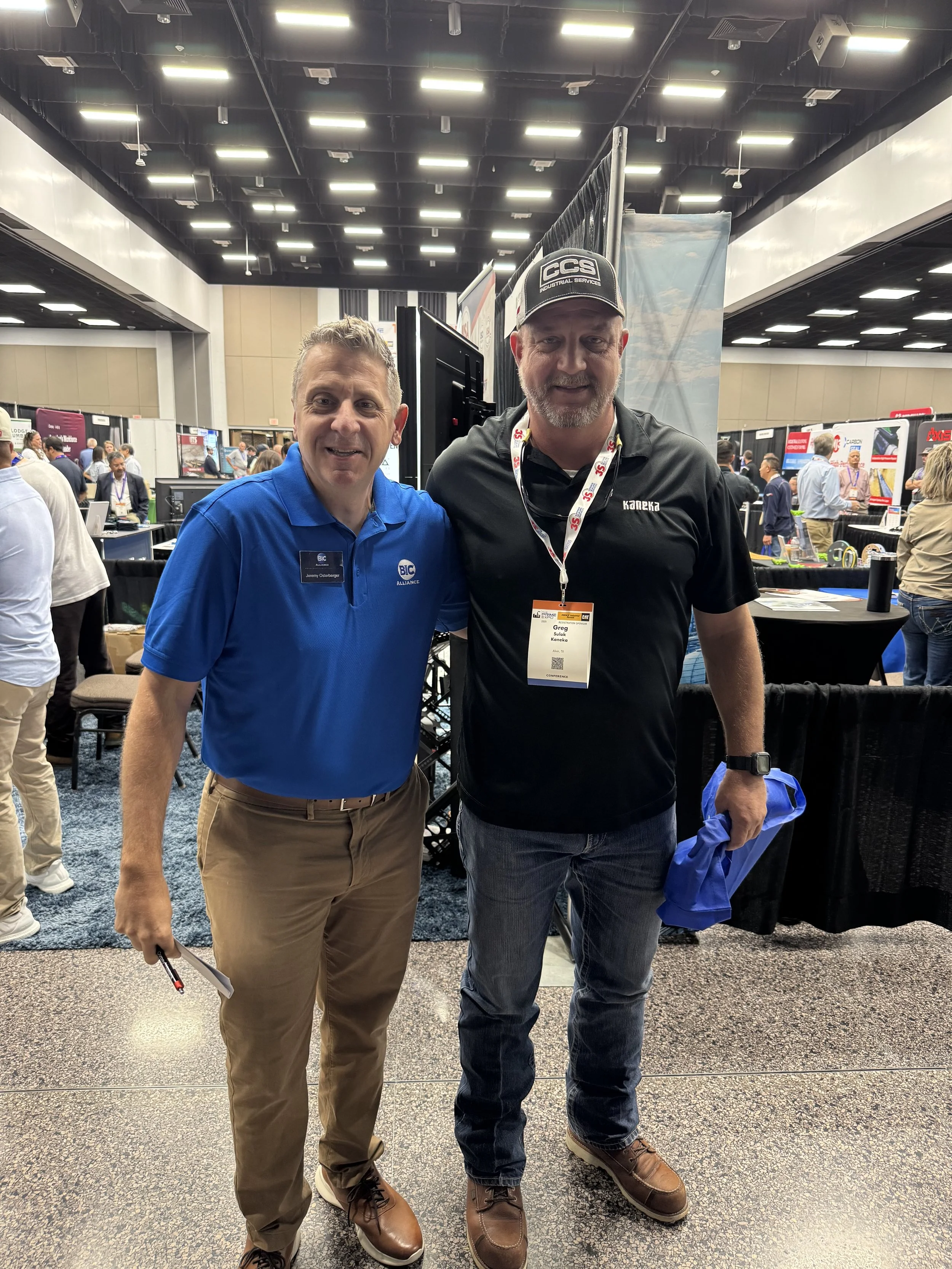 Two men standing together at a conference or trade show, smiling for the photo. One man is wearing a blue polo shirt with an ID badge, and the other is wearing a black shirt with a cap, also with a badge. The background shows other attendees, booths,