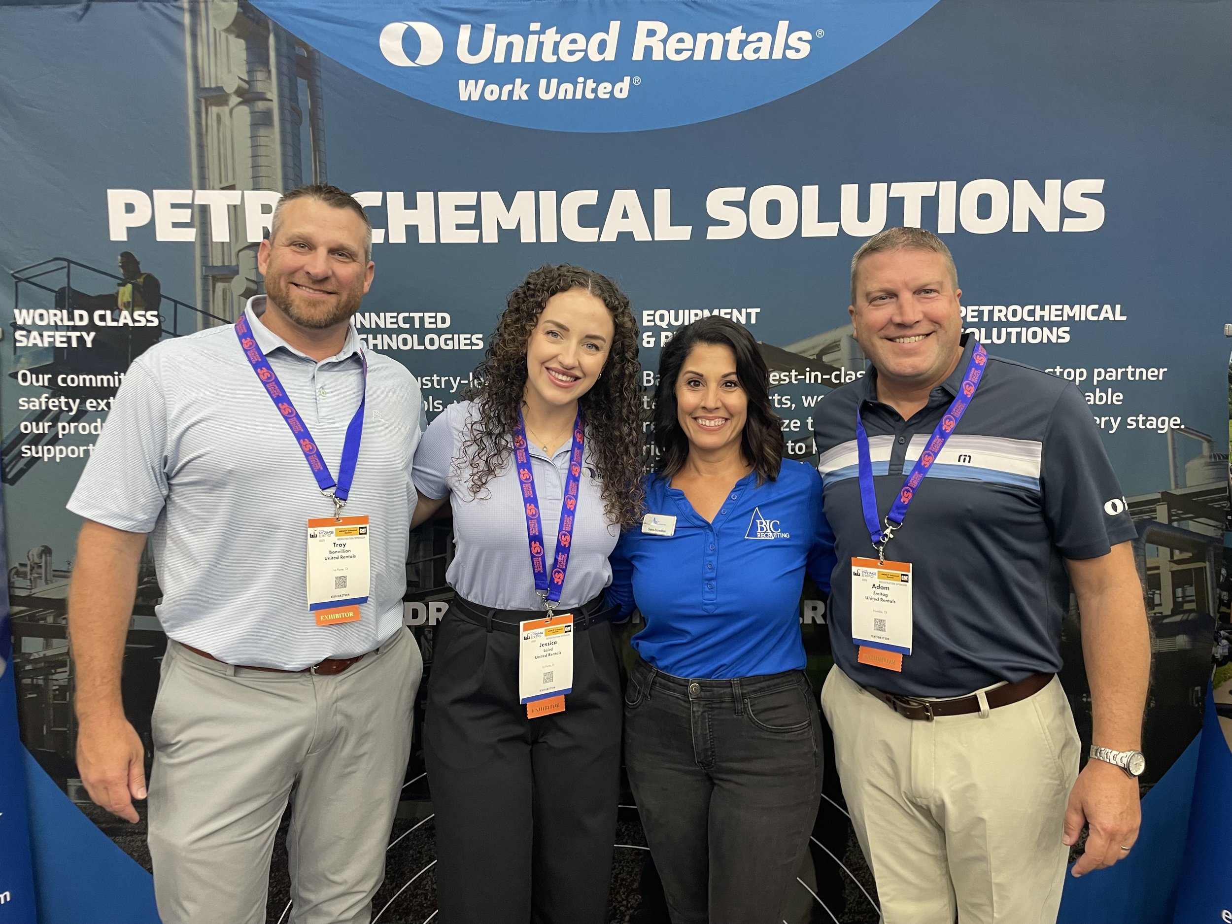 Four people standing in front of a banner at a conference, smiling and wearing name badges, with a blue background showing industrial equipment and the United Rentals logo.