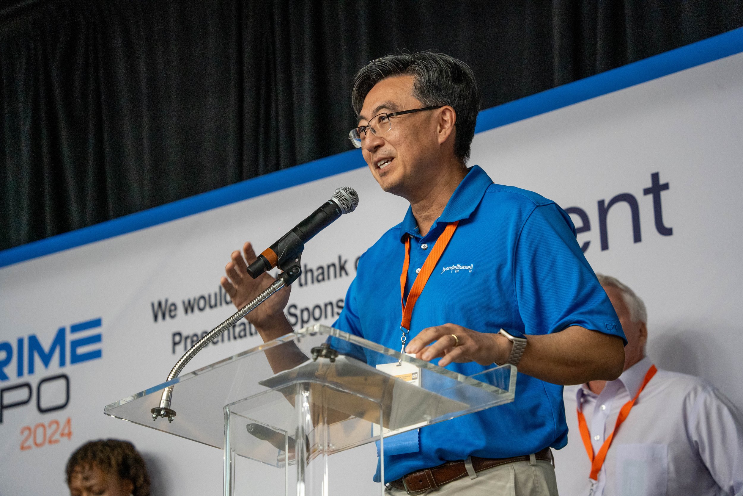 A man with glasses and dark hair, wearing a blue polo shirt, stands behind a clear lectern with a microphone, speaking at an event. There is a large banner behind him with text and logos, including the words 'PRIME' and '2024'.