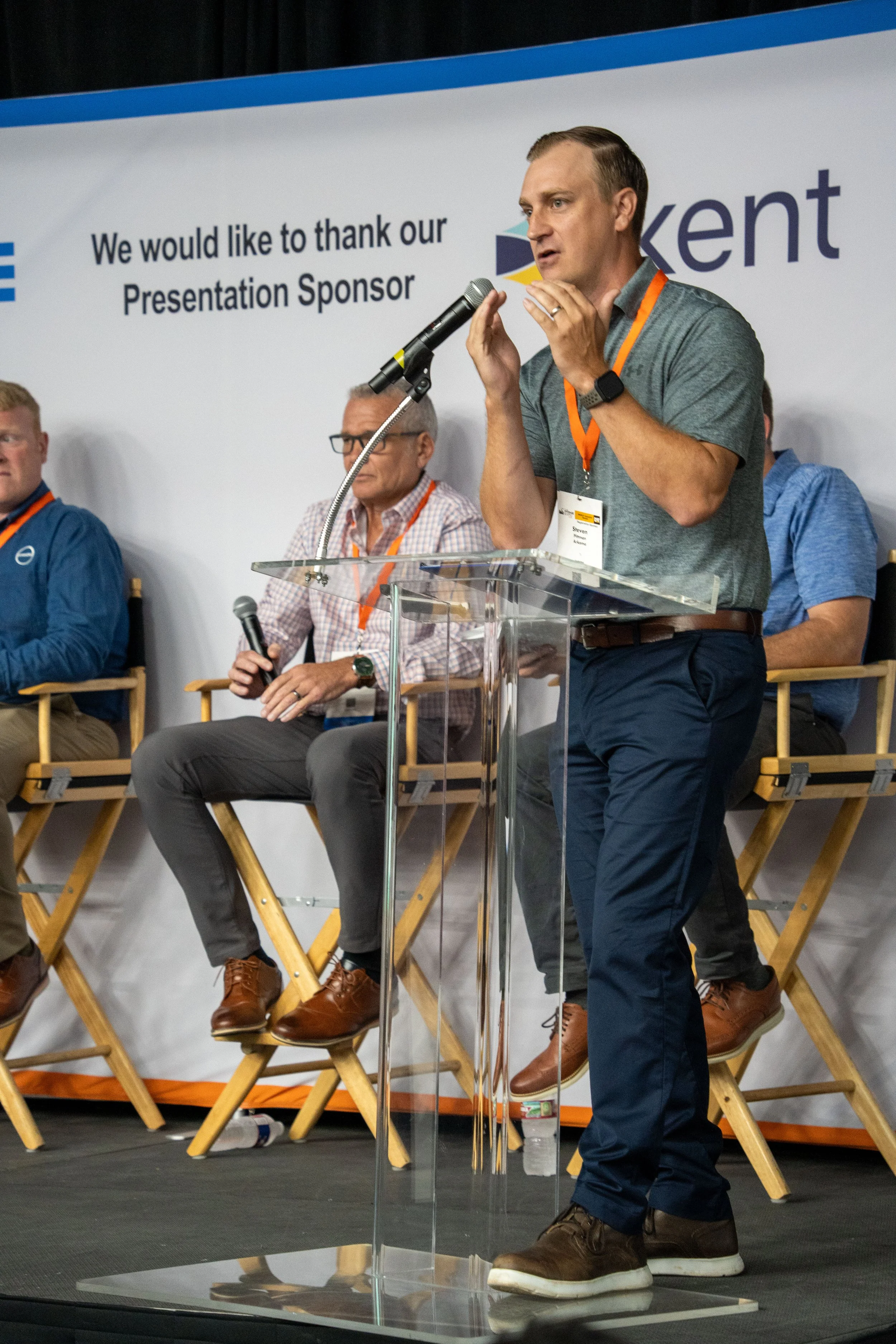 A man speaking at a podium during a conference, with other seated panelists behind him, a large banner with text and logos in the background.