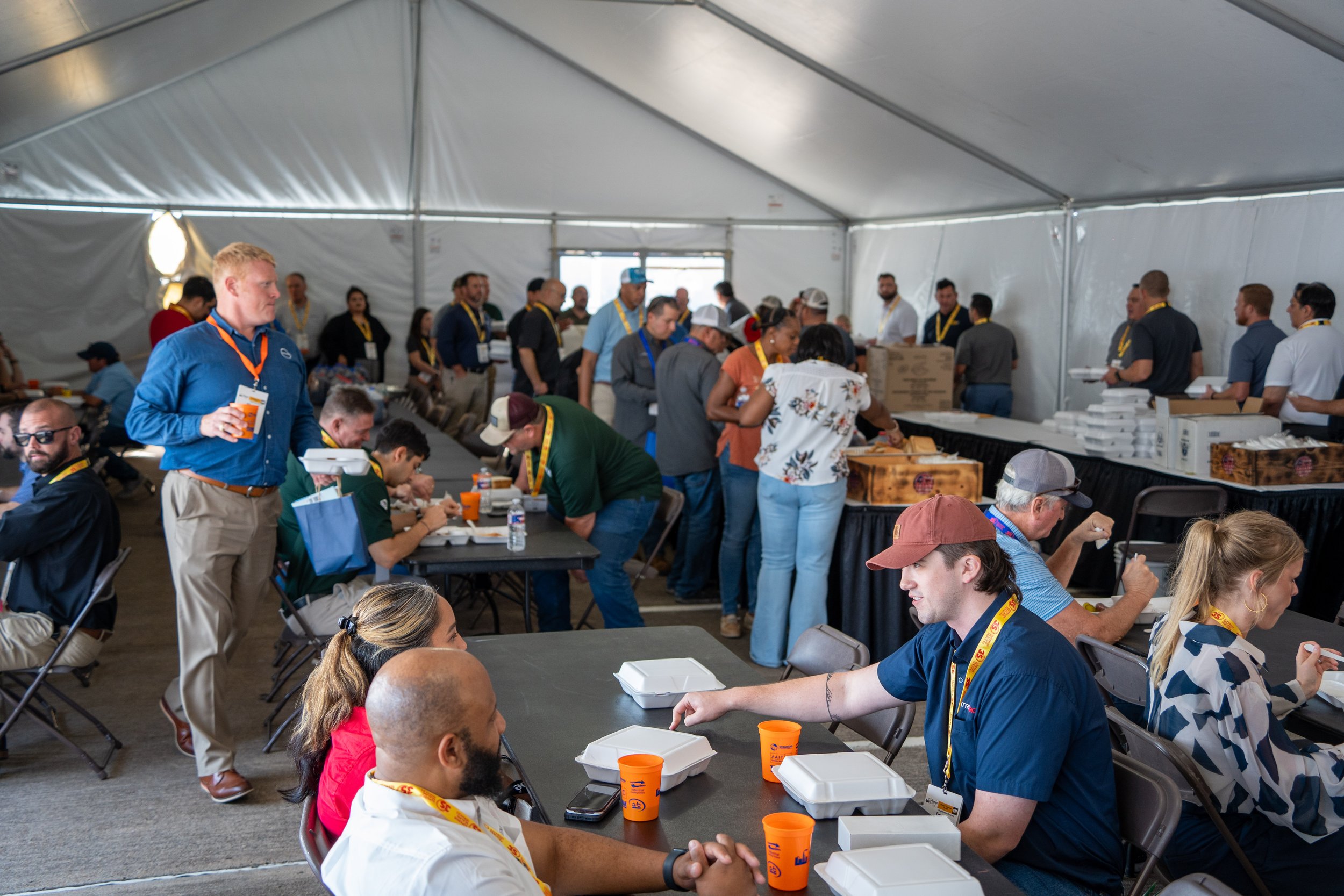 People in a large tent eating and waiting in line for food at a food service event, with many serving dishes and boxes of food on tables.