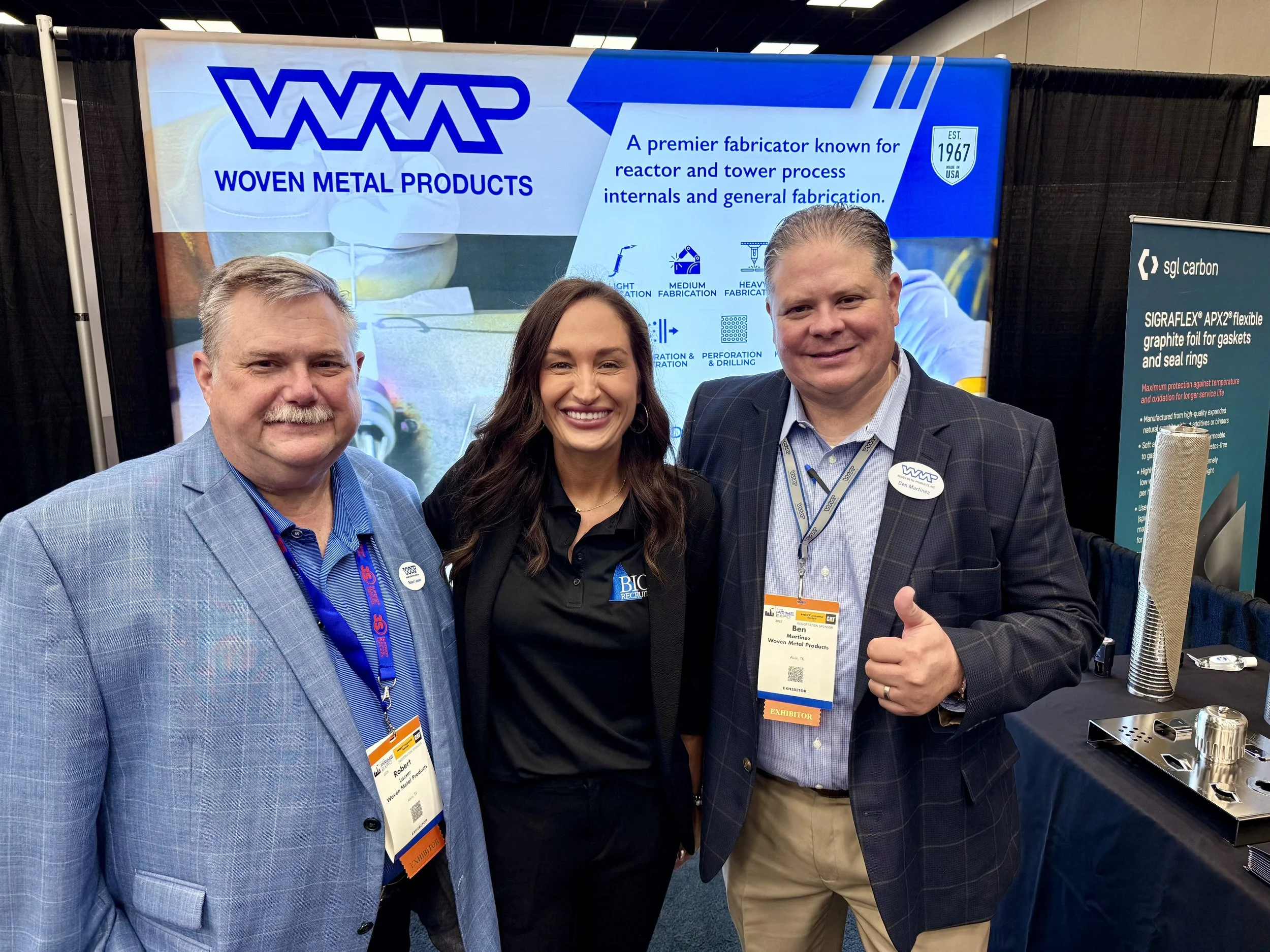 Three people standing in front of Woven Metal Products exhibit at a trade show, smiling and posing for the photo.