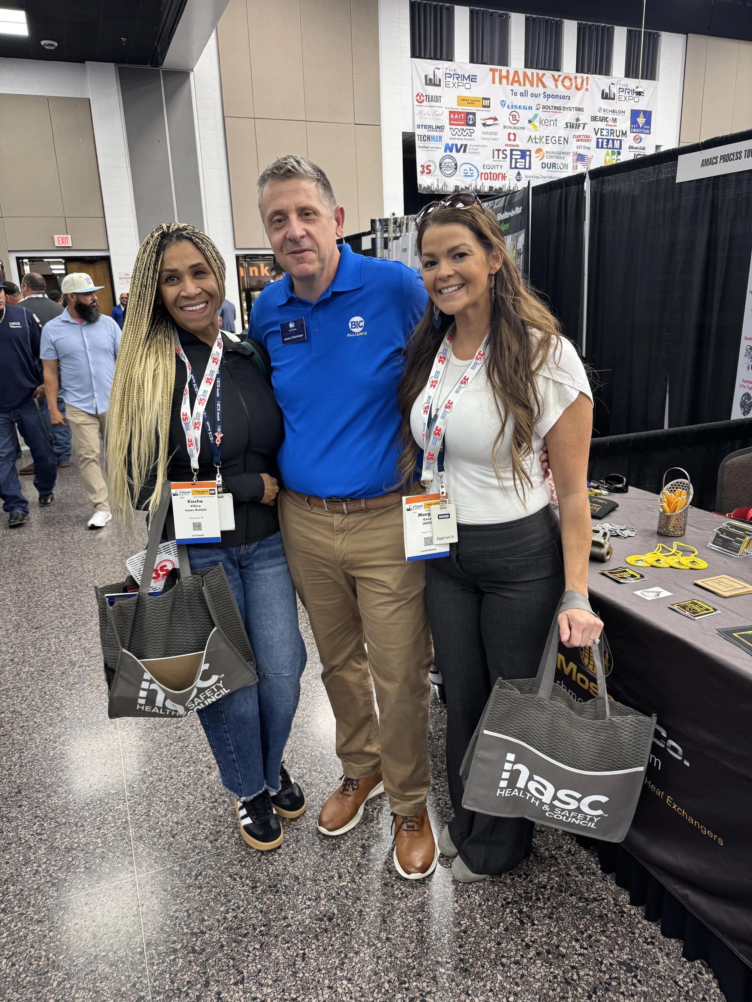 Three people standing together at a conference, smiling for the camera. They are wearing name badges on lanyards, and two are holding tote bags with "HASC" written on them. The background shows a large thank you banner with many sponsor logos.