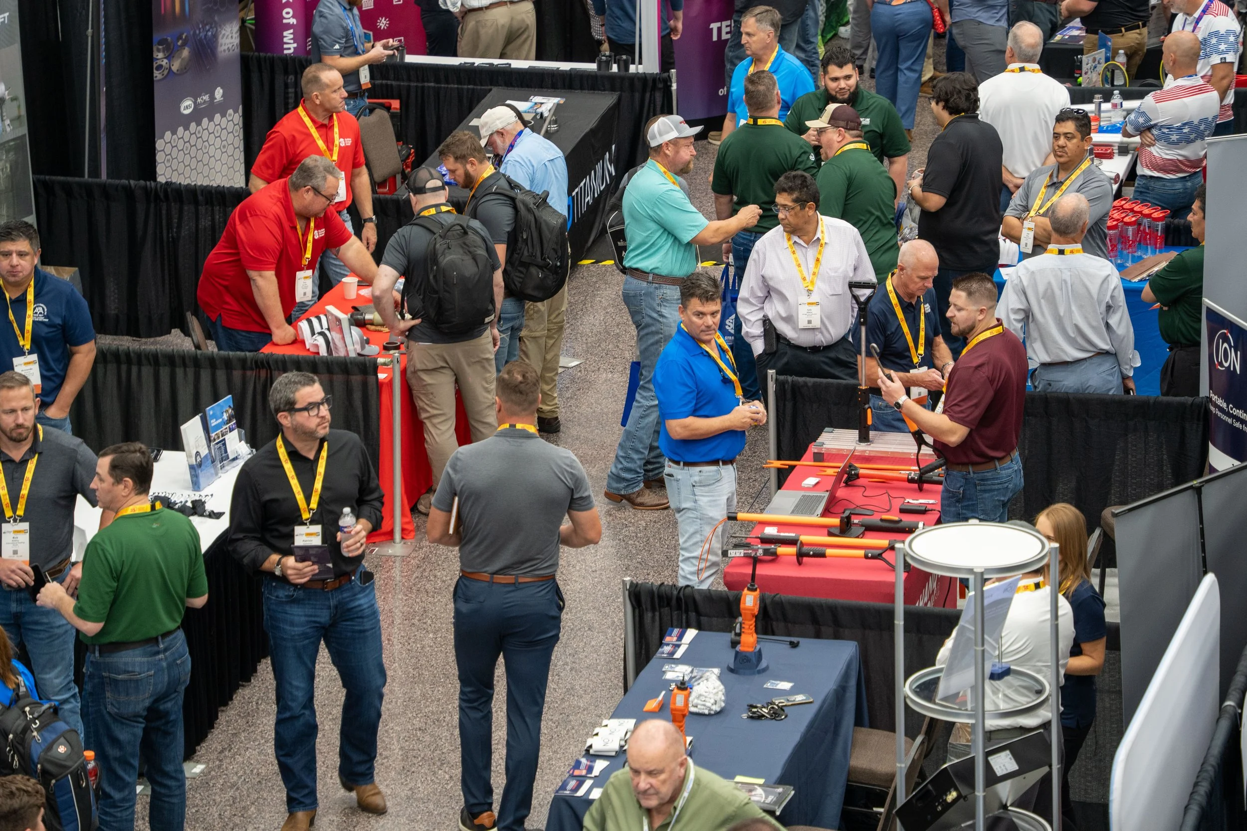 Trade show with booths and attendees examining products, including a man demonstrating a tool to another person at a table with orange equipment.