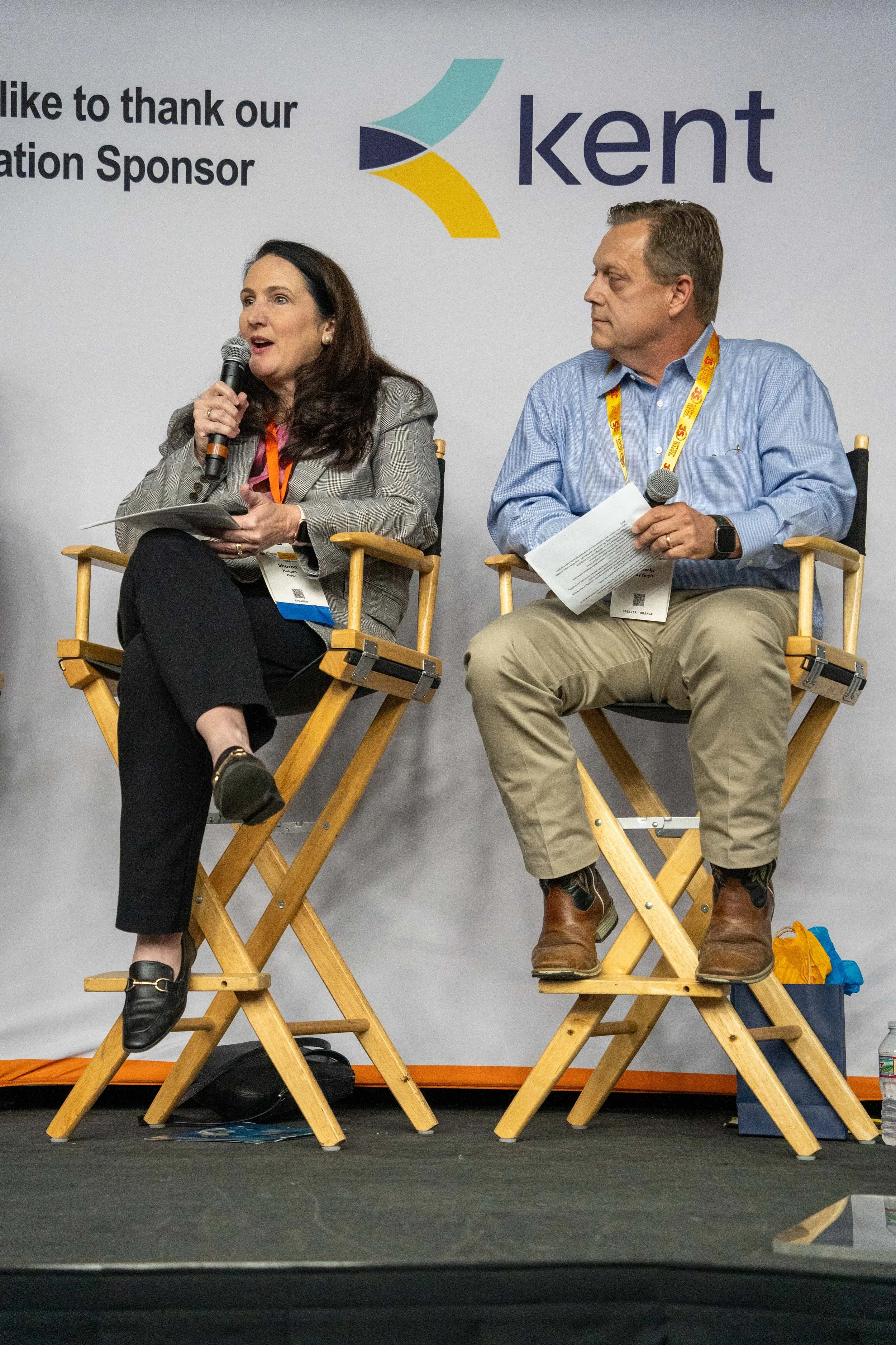 Two people sitting on wooden director chairs on a stage at a conference, with a white backdrop displaying a logo and partial text. The woman on the left is speaking into a microphone, and the man on the right is looking at her, holding papers and a m