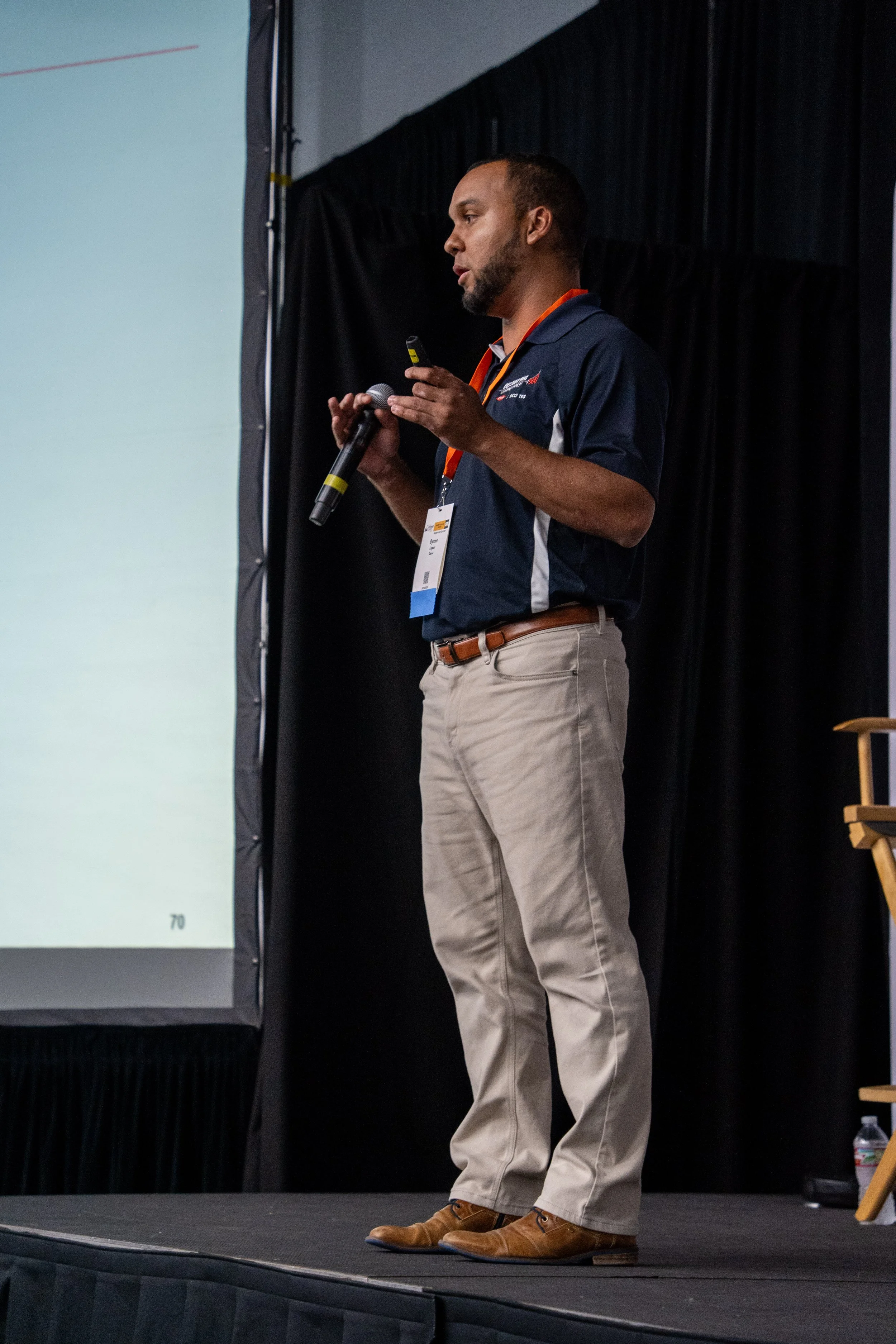 A man giving a presentation on a stage with a black curtain backdrop, holding a microphone and a clicker, wearing a navy polo shirt with white accents and khaki pants.