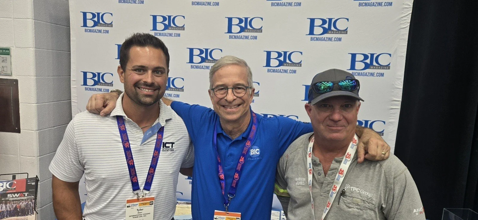 Three men standing together at a conference or event, smiling, with a backdrop displaying BIC Magazine logos. The man on the left wears a white and gray striped polo shirt and a lanyard. The man in the middle wears glasses and a blue shirt. The man o