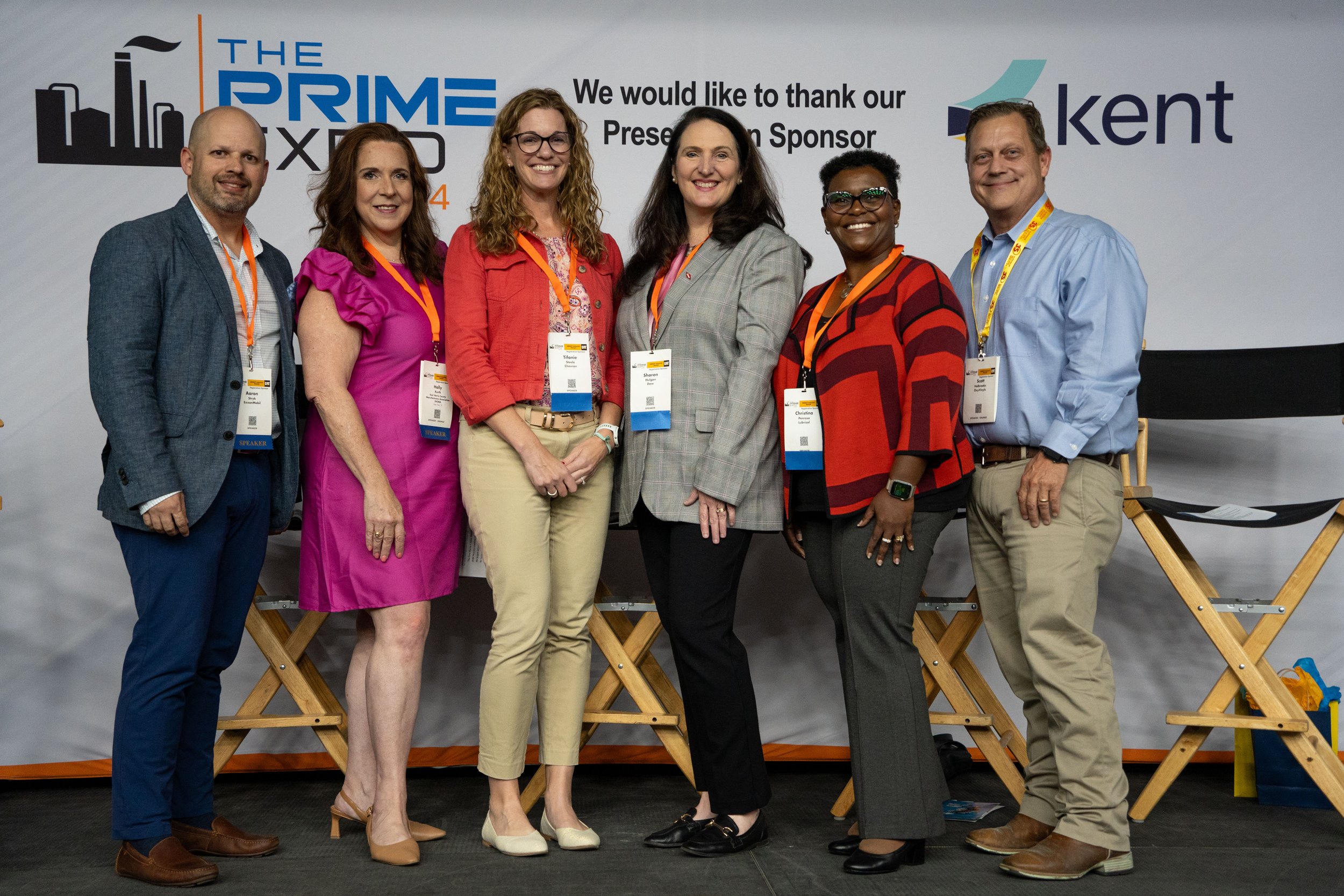 A group of seven people standing in front of a conference backdrop, smiling for the photo. The backdrop features the logos of 'The Prime', 'Kent', and a slogan thanking the sponsor. The individuals are wearing conference name badges on orange lanyard