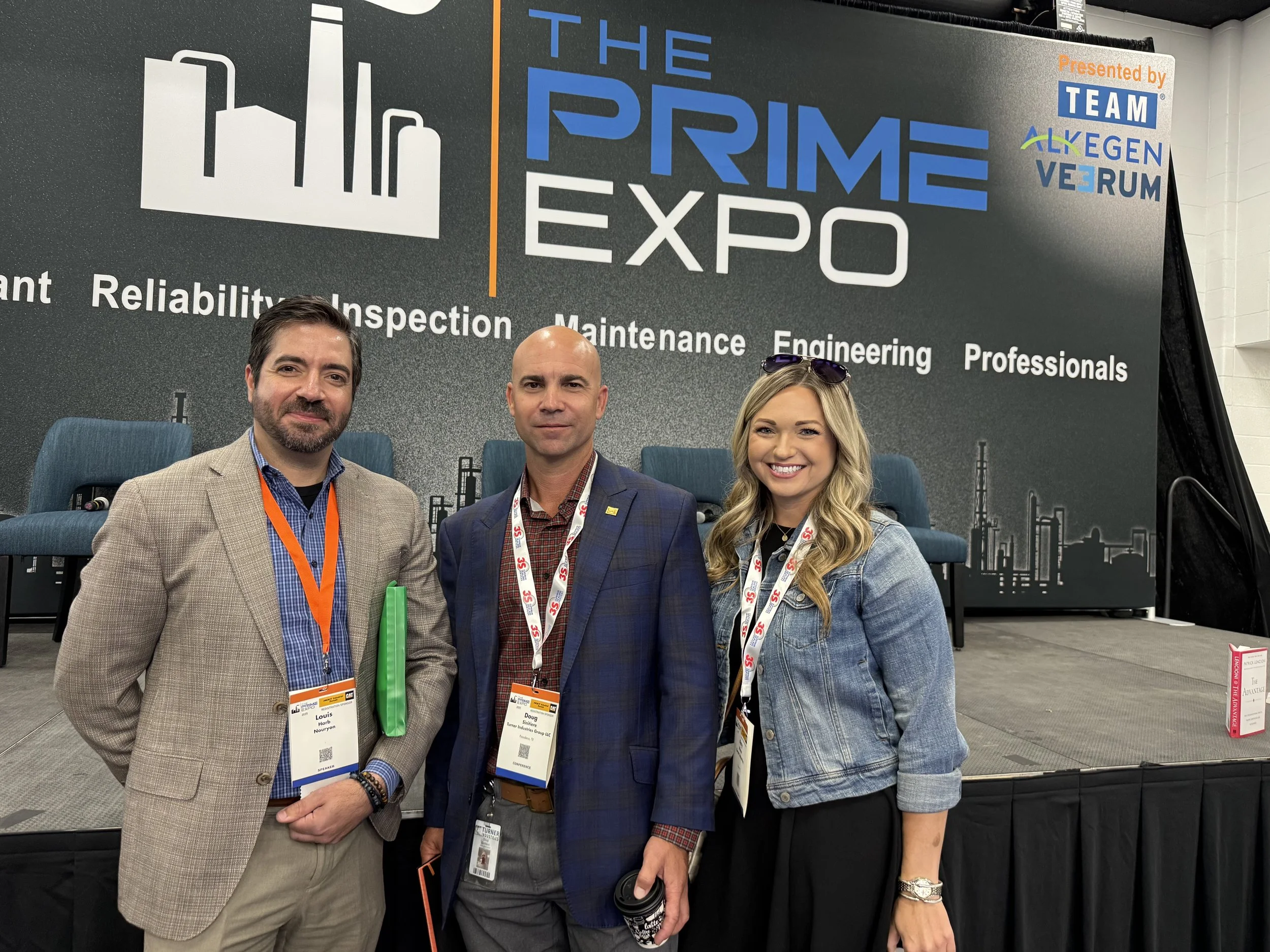Three professionals standing in front of a stage with a backdrop at the Prime Expo. The backdrop displays the event title, topics like reliability, inspection, maintenance, engineering, professionals, and logos, including Team AL-KEGEN VE-RUM. The th