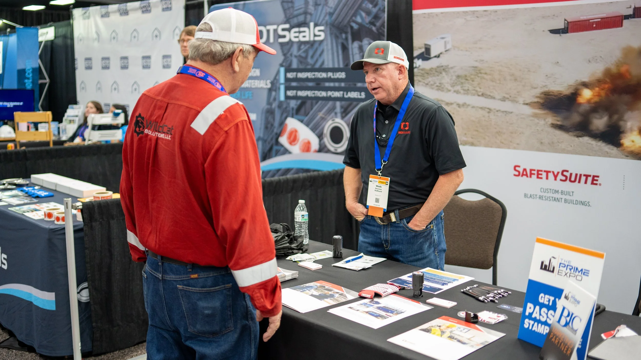Two men talking at an exhibition booth with safety and construction branding; one man wearing a red jacket and the other in a black polo shirt with event badges.