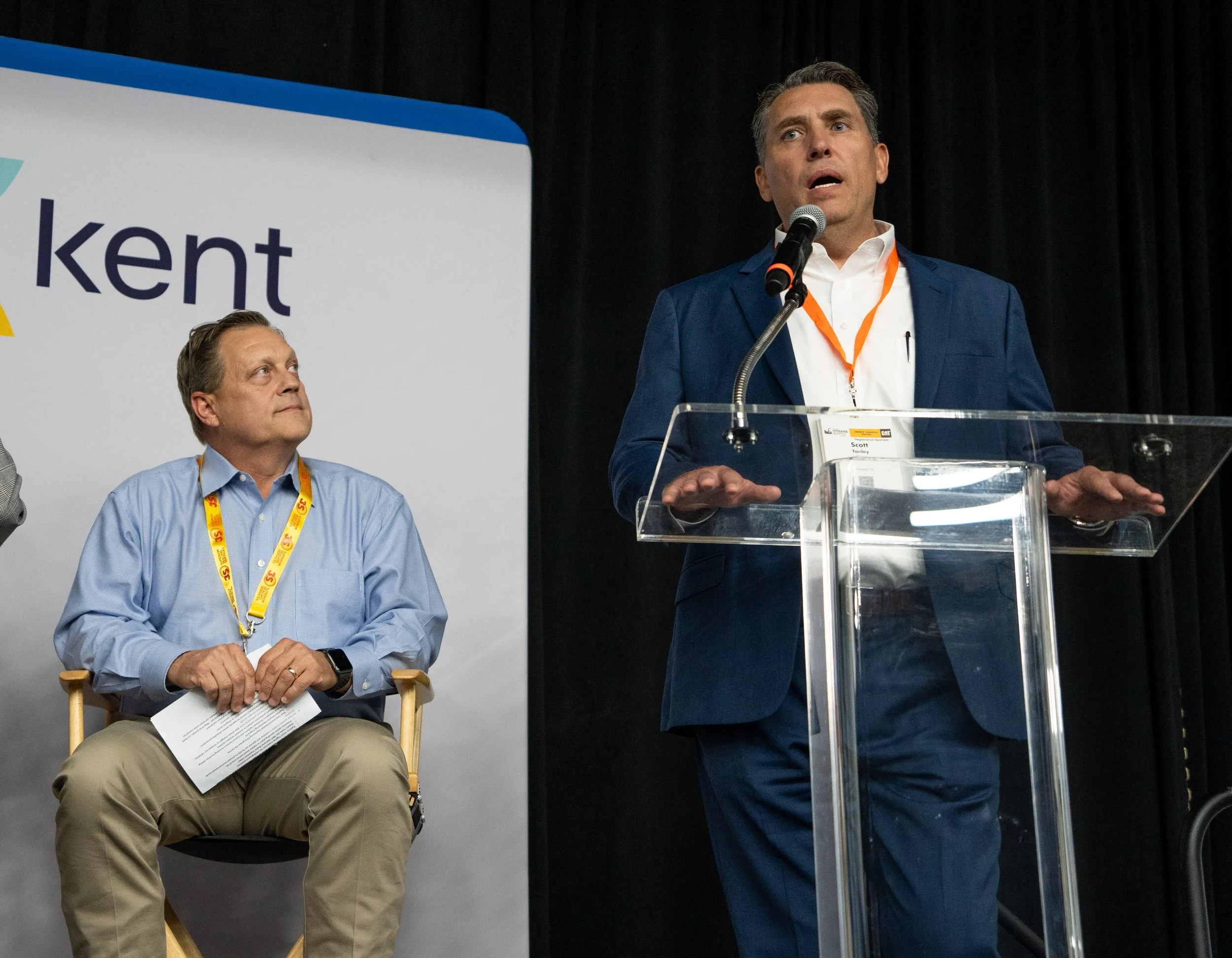 A man in a blue suit is speaking at a podium with a microphone, while another man in a light blue shirt sits nearby, looking up at him. The background features a sign with part of the word "Kent."