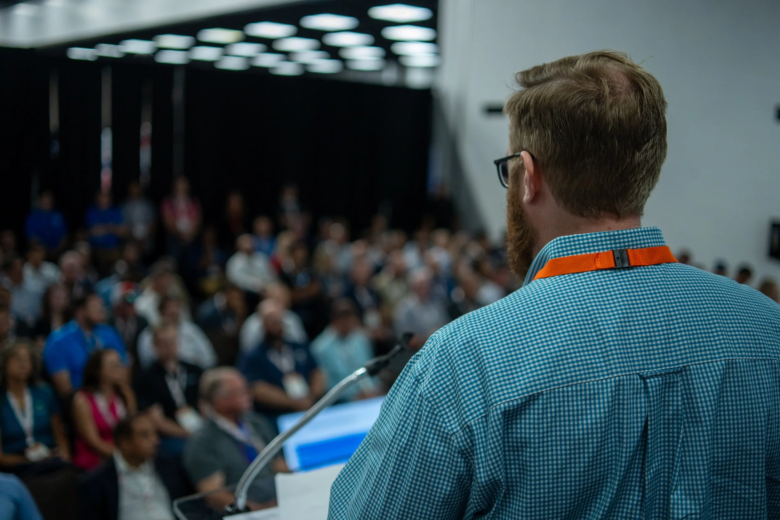 A man with glasses and a beard stands at a podium, speaking to a large, seated audience in a conference or seminar room.