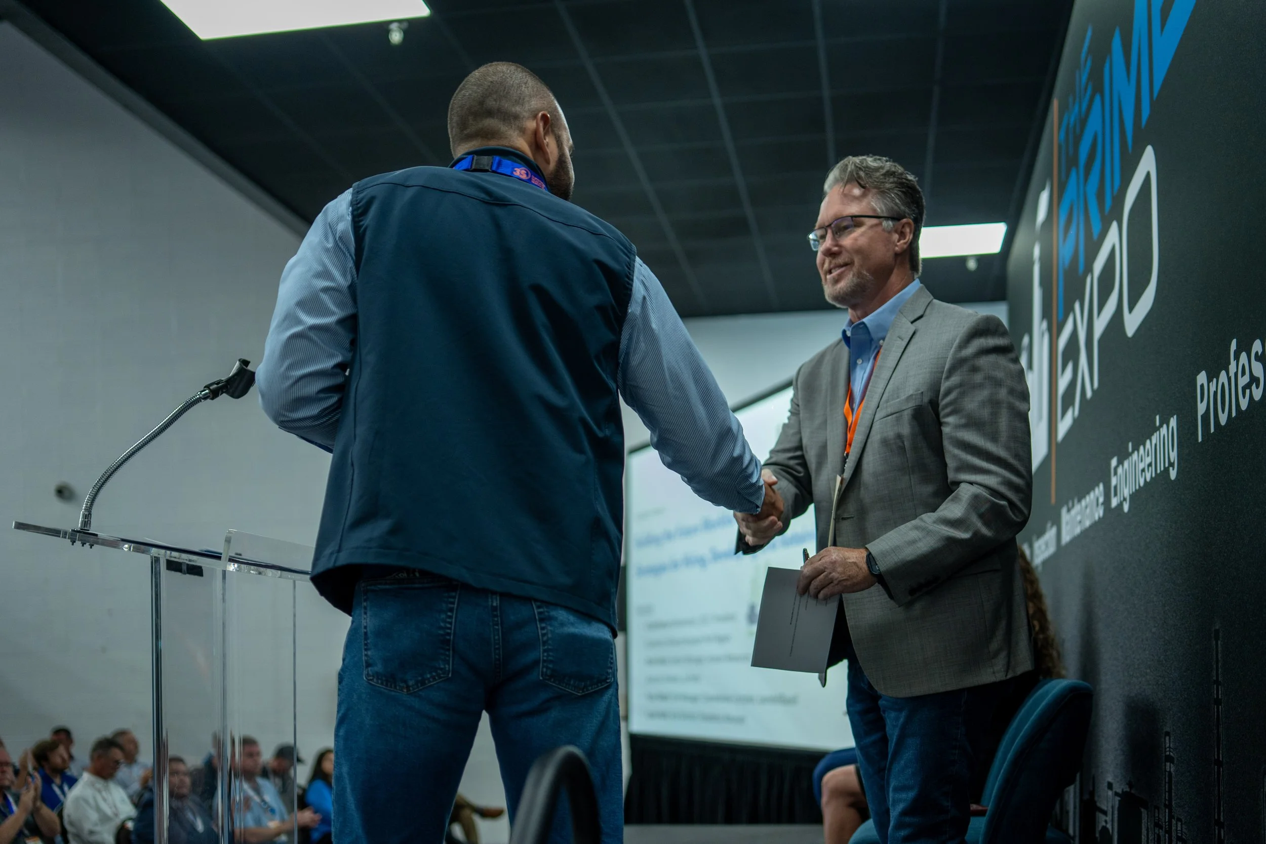 Two men shake hands on stage during a conference, with an audience watching. One man wears a gray blazer and glasses, and the other wears a navy vest and jeans.