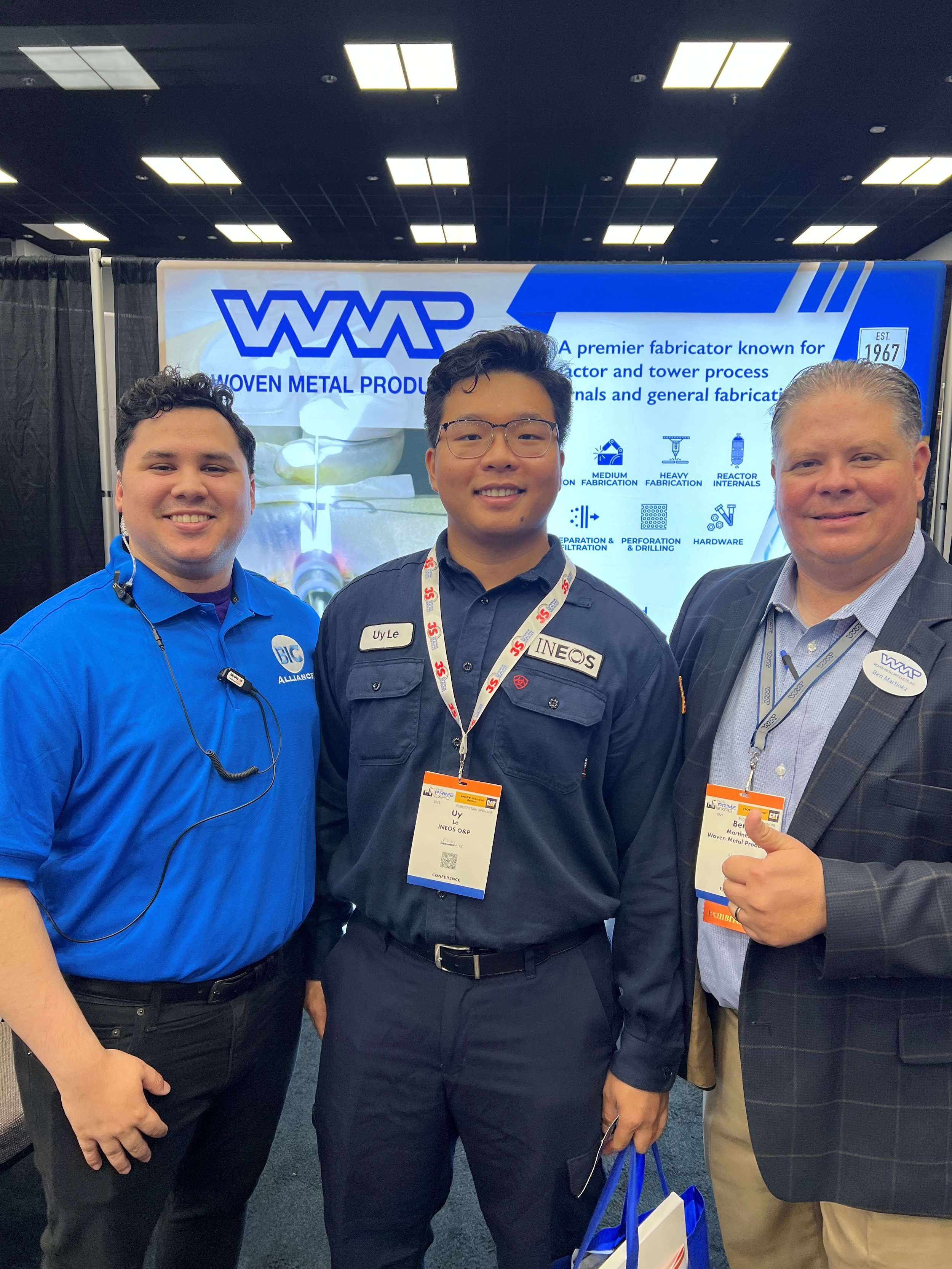 Three men standing together at a trade show or conference, smiling and posing for the camera. Behind them is a large display screen showing information about woven metal production, with a blue and white background and text.