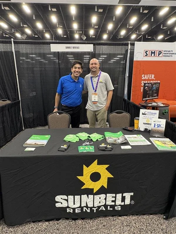 Two men standing behind a table with promotional materials at an exhibit booth for Sunbelt Rentals inside a convention center. The table has a black cover with the Sunbelt Rentals logo, and the booth has a black backdrop with signs for Sunbelt Rental