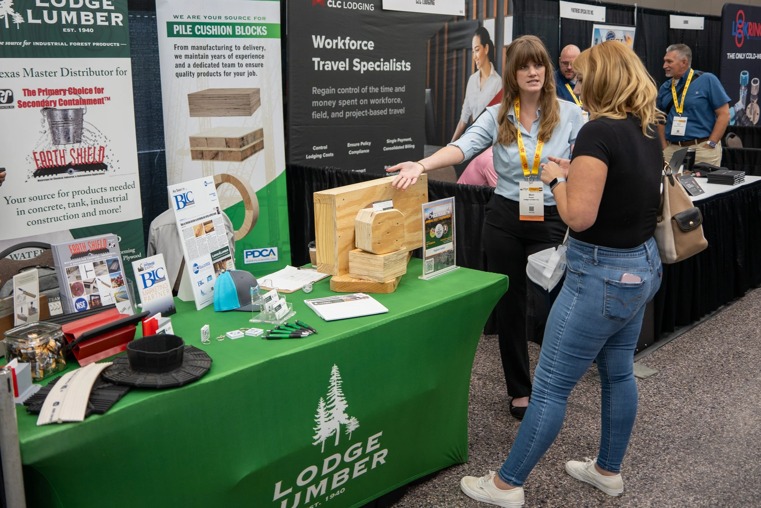 A woman with brown hair and black shirt talking with a woman with brown hair and a light blue shirt at a trade show booth for Lodge Lumber, with wooden samples and promotional materials on the table.