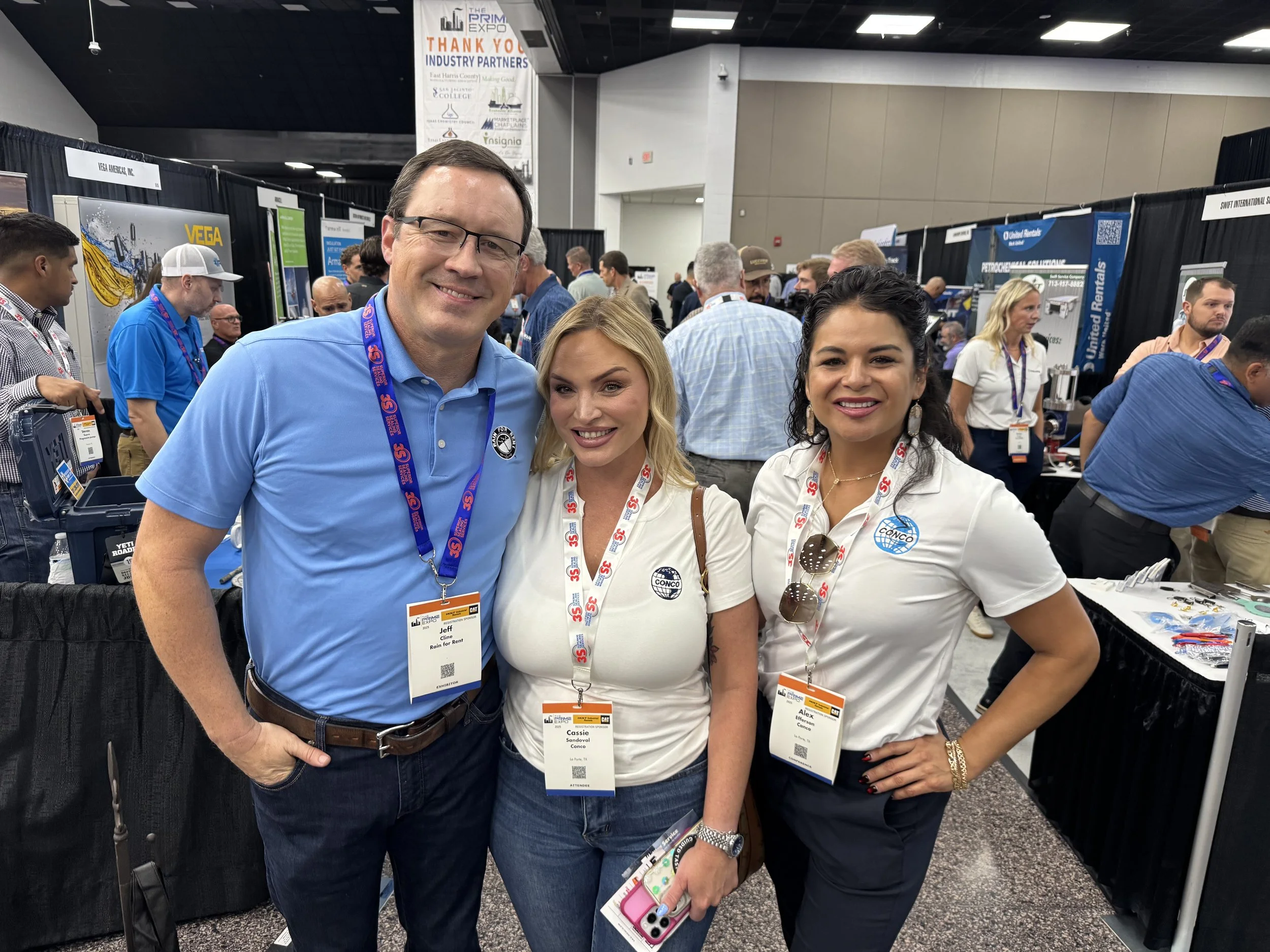 Three people smiling at a conference, wearing conference badges and lanyards, standing in front of exhibition booths with various displays and attendees in the background.