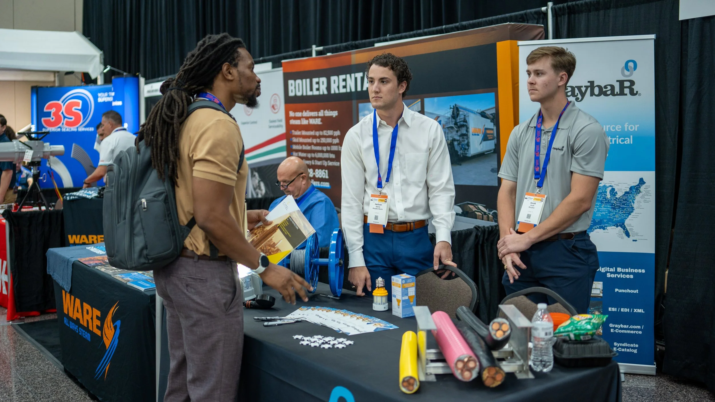 Two men in business attire talking to a visitor at a trade show booth, with various electrical and lighting products on the table, and banners advertising boiler rental services and electrical resources in the background.