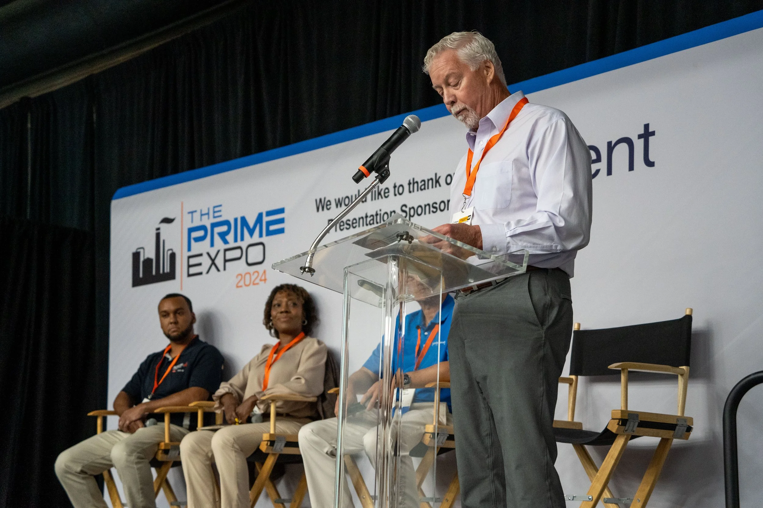 A man with white hair and a white shirt speaking at a podium during the Prime Expo 2024 event, with three people seated behind him on stage.