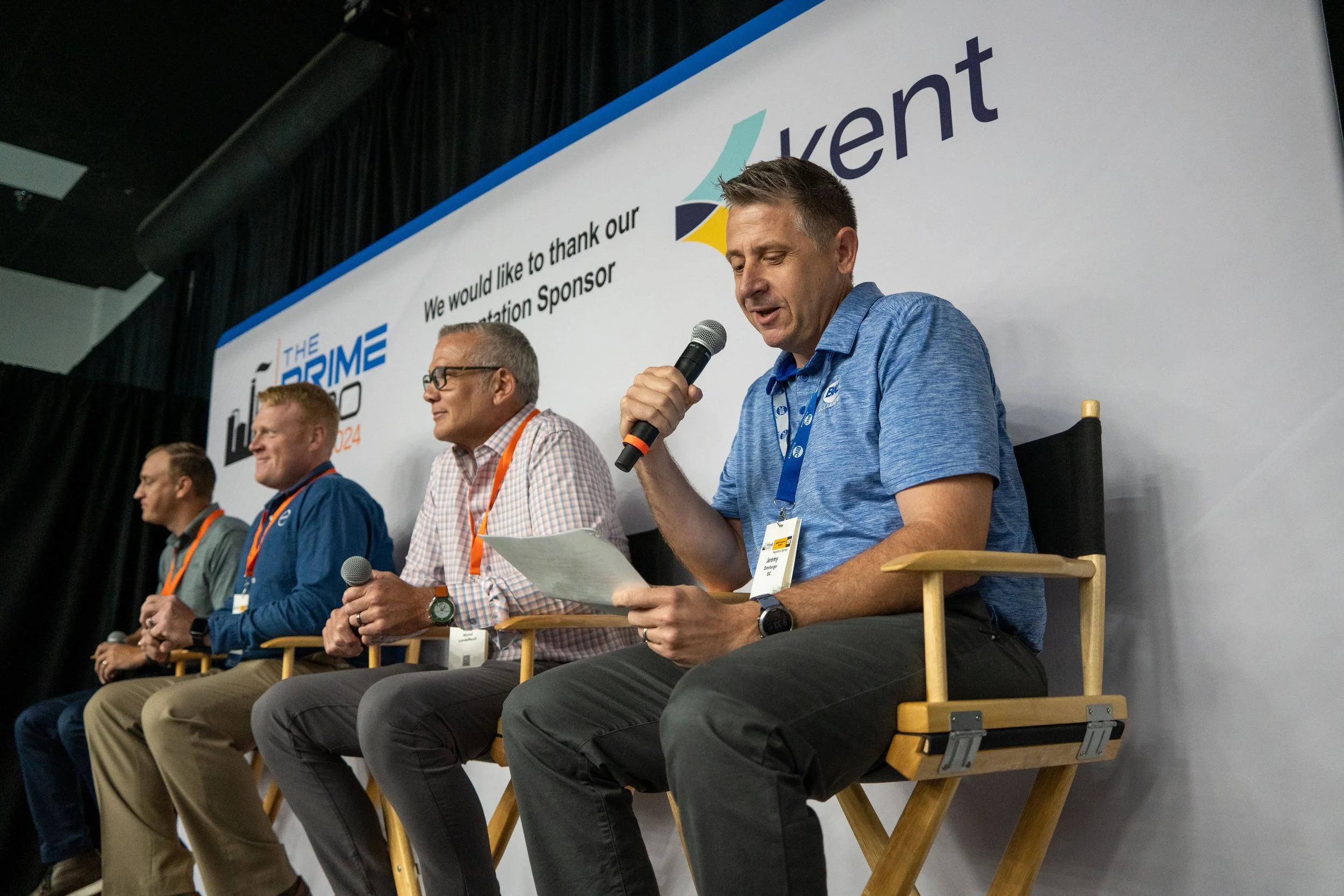 Four men sitting on a stage in front of a white backdrop, with one man speaking into a microphone and holding a paper.