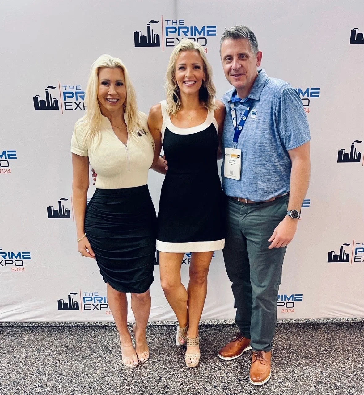 Three people standing together at an event called The Prime Expo 2024, posing in front of a backdrop with the event's logo. The person on the left is a woman with long blonde hair wearing a cream-colored top with a zipper and a black ruched skirt. Th