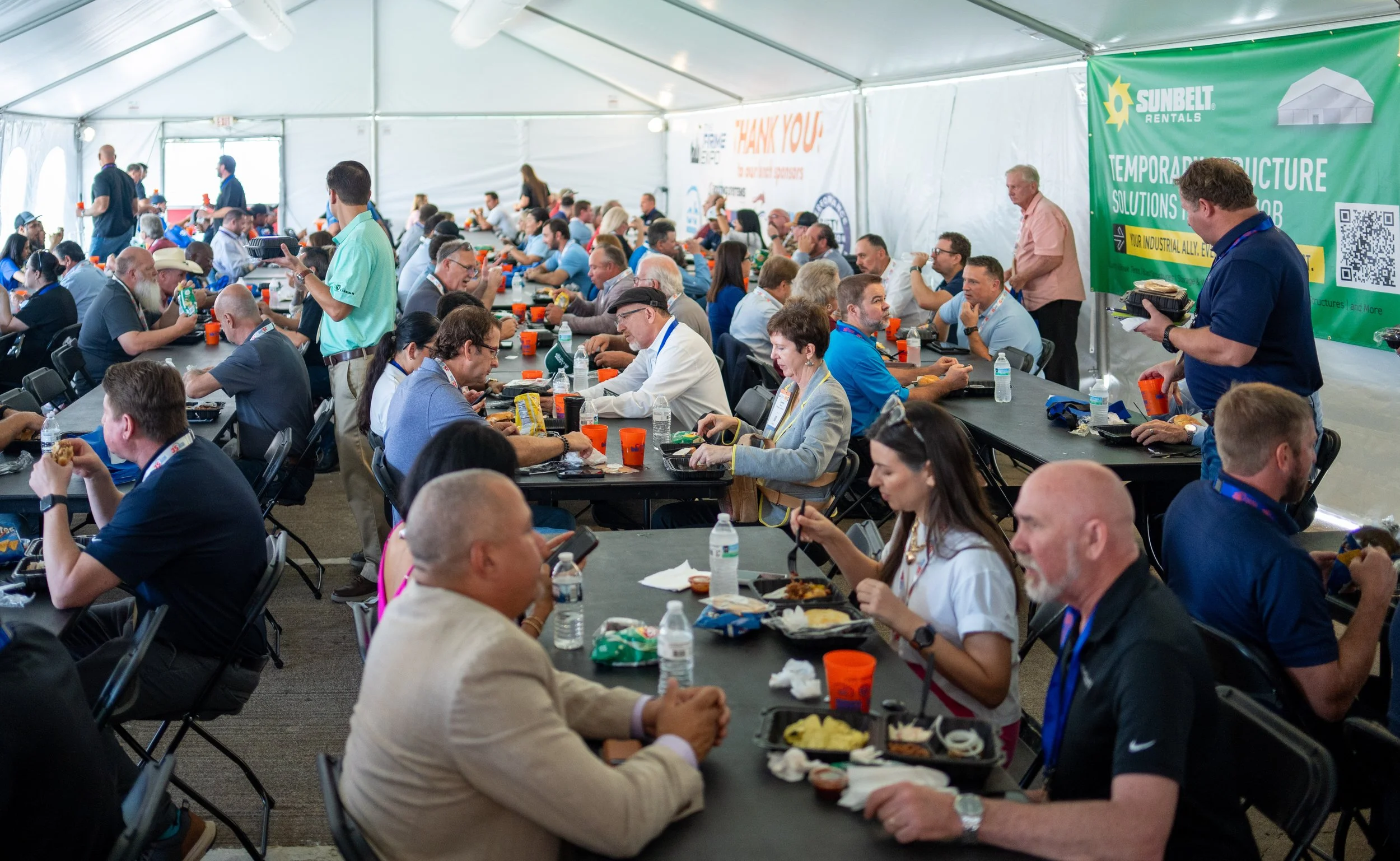 People seated at long tables in a large white event tent, eating lunch. Several individuals are engaged in conversations or using their phones. There are banners on the walls advertising rental services and construction solutions.