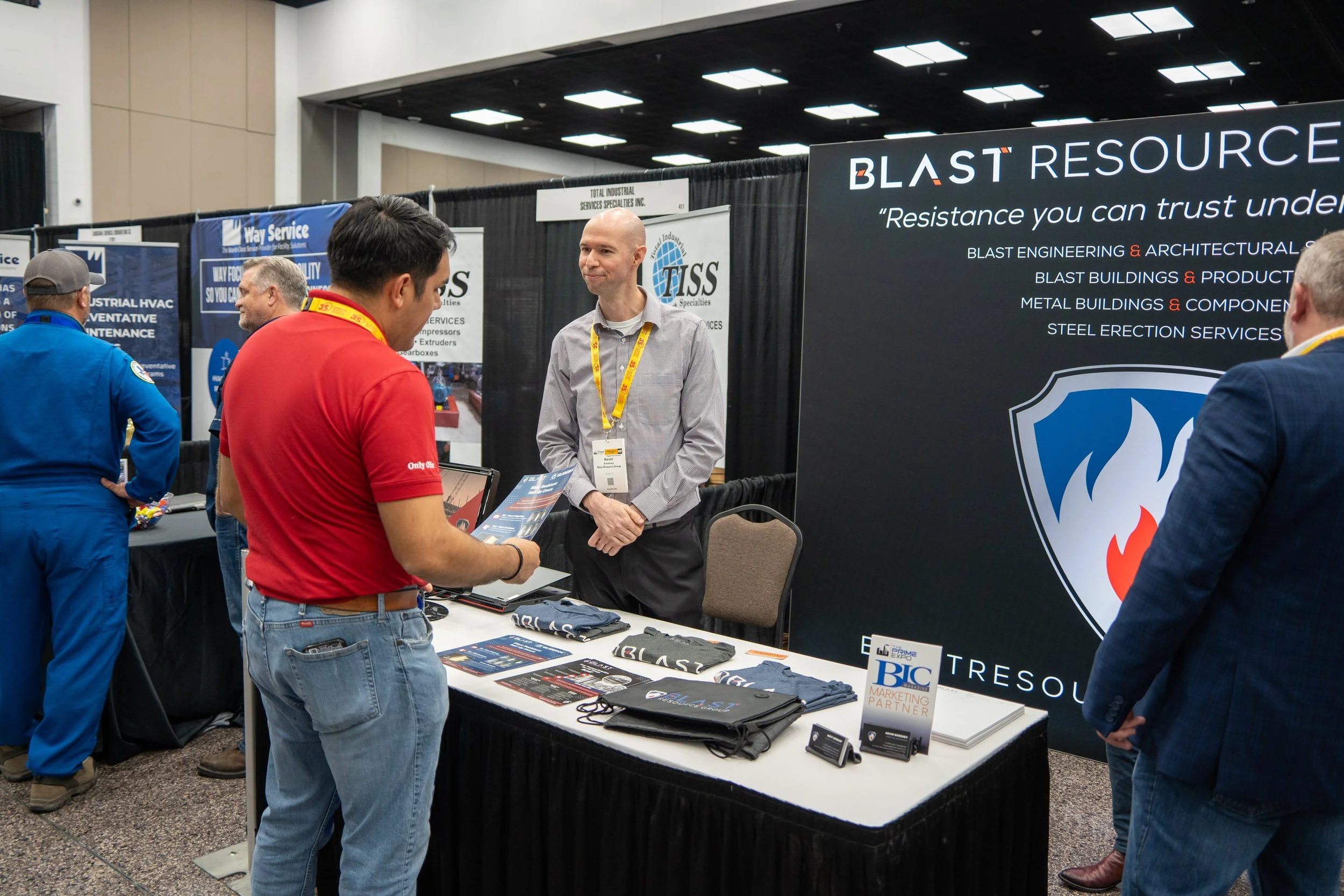 A trade show booth showcasing a company called BLAST RESOURCES with a large black banner displaying their services, including steel erection and architectural services. A man with a gray shirt and yellow lanyard is talking to a visitor wearing a red 