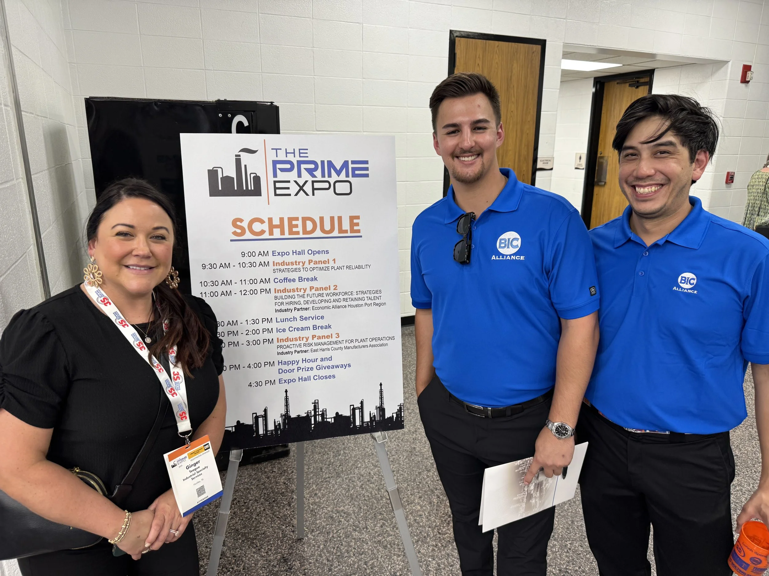 Three people standing together at a conference near a schedule sign. The woman on the left is wearing a black dress with a name badge, the two men on the right are in matching blue shirts with a company logo, and they are smiling at the camera.