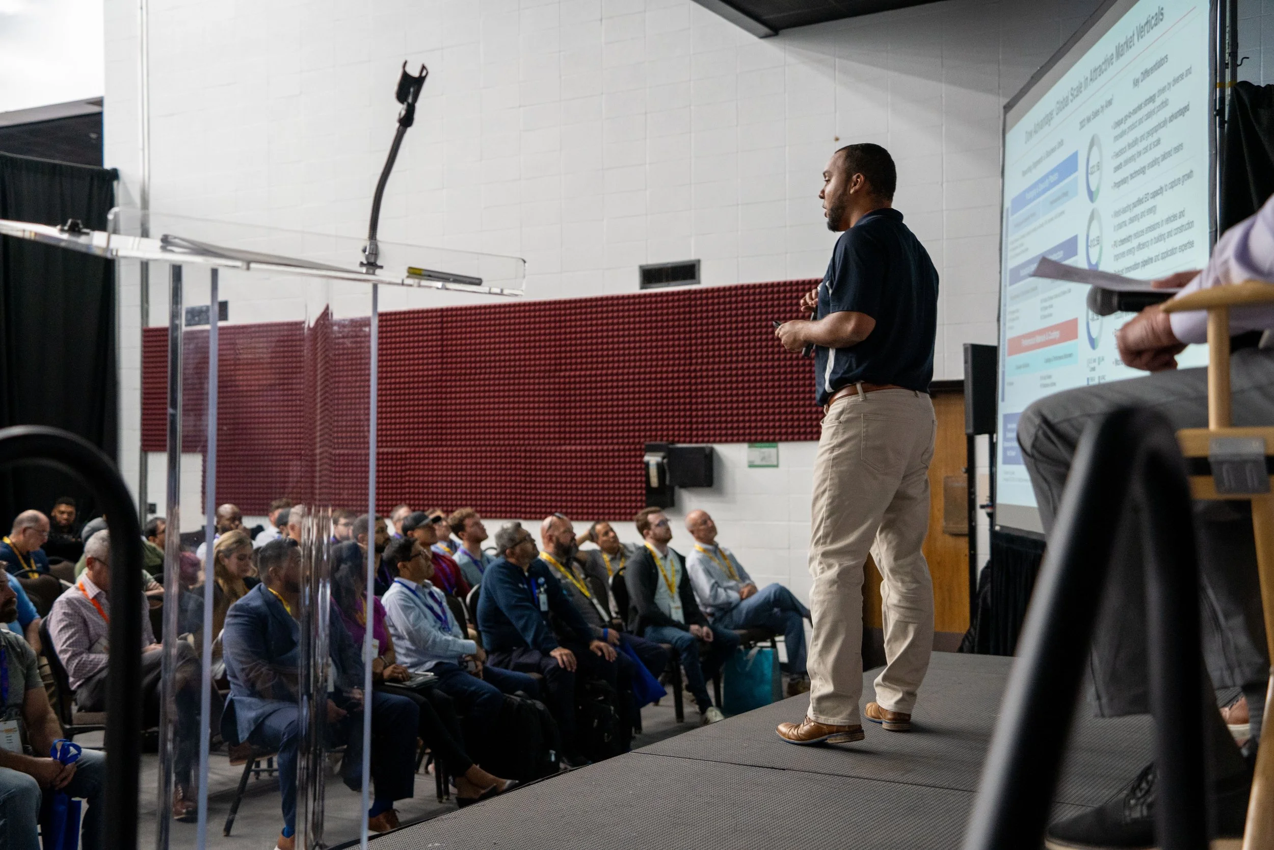 A man is giving a presentation in front of a seated audience at a conference or seminar, with a large display screen showing informational slides.