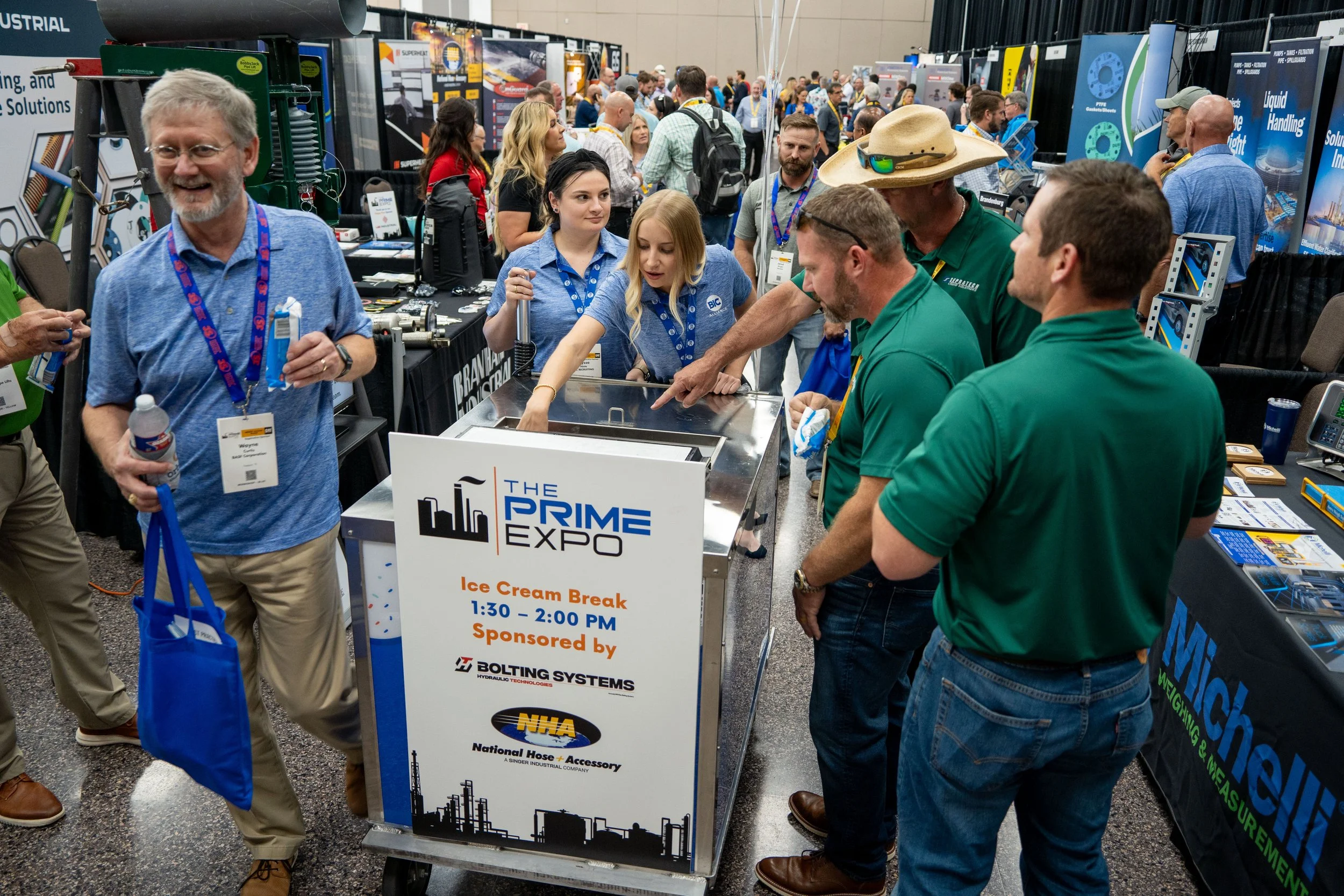 Attendees at the PRIME EXPO examining an ice cream machine during the ice cream break event, sponsored by Bolting Systems and NHA, with a crowd in the background at a trade show.