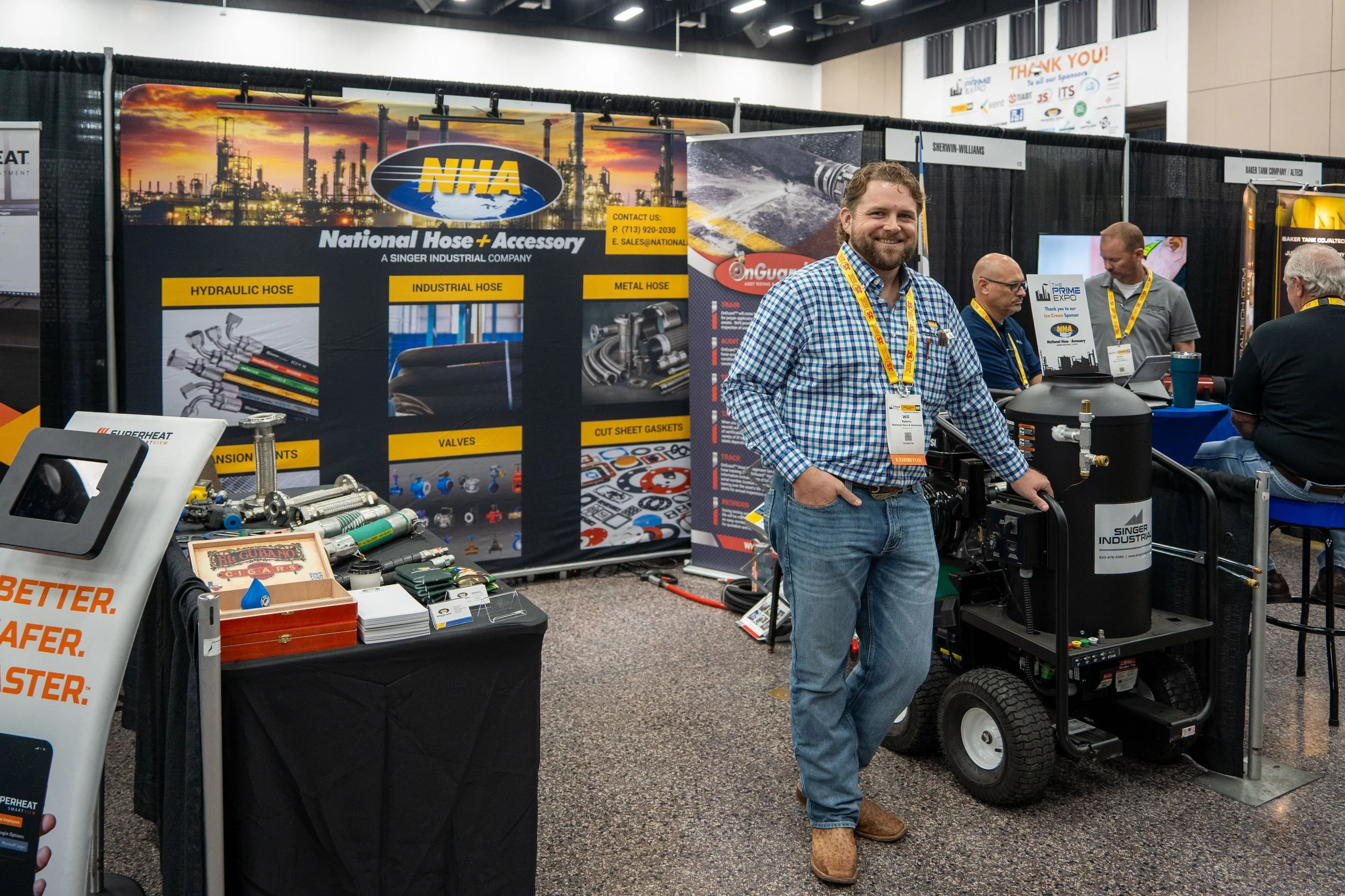 Man smiling at a trade show booth with industrial hoses and fittings on display, and a banner reading 'NHA - National Hose + Accessory' in the background.