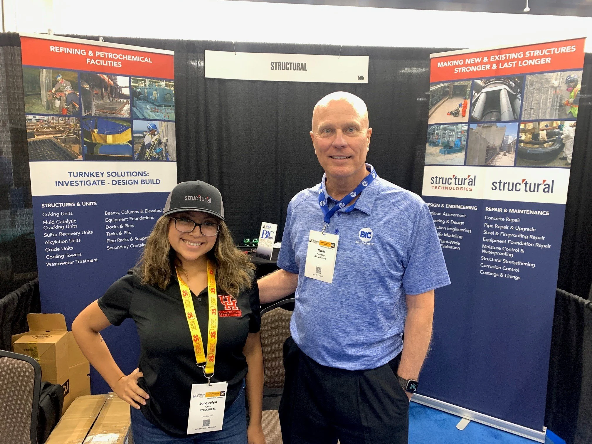 Two people smiling at a trade show booth with blue and red banners. The woman is wearing glasses, a gray hat, a black polo shirt with a red logo, and a yellow lanyard. The man is wearing a blue polo shirt, a wristwatch, and a lanyard. The banners beh