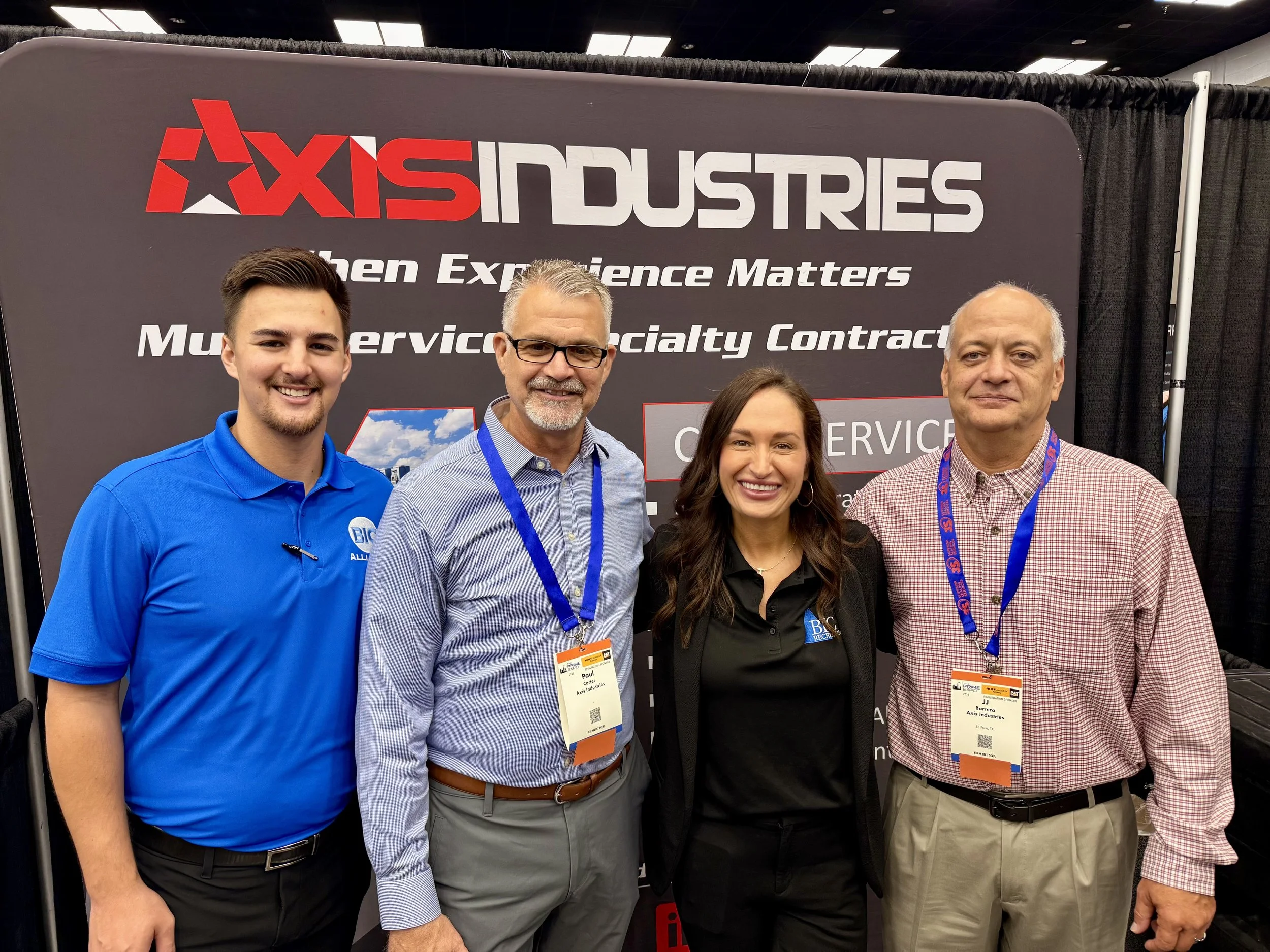 Four people standing in front of a black and red sign at an event. The sign reads "AXIS INDUSTRIES" and "Other Experience Matters" and mentions "Multiple Service & Contract". The group includes two men and one woman smiling at the camera, all wearing