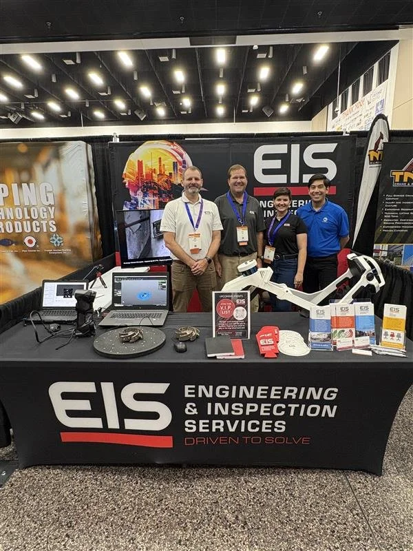 A booth at a conference or trade show for EIS Engineering & Inspection Services, featuring a table with computers and equipment, and four smiling people standing behind it. The booth has banners and brochures promoting their services.